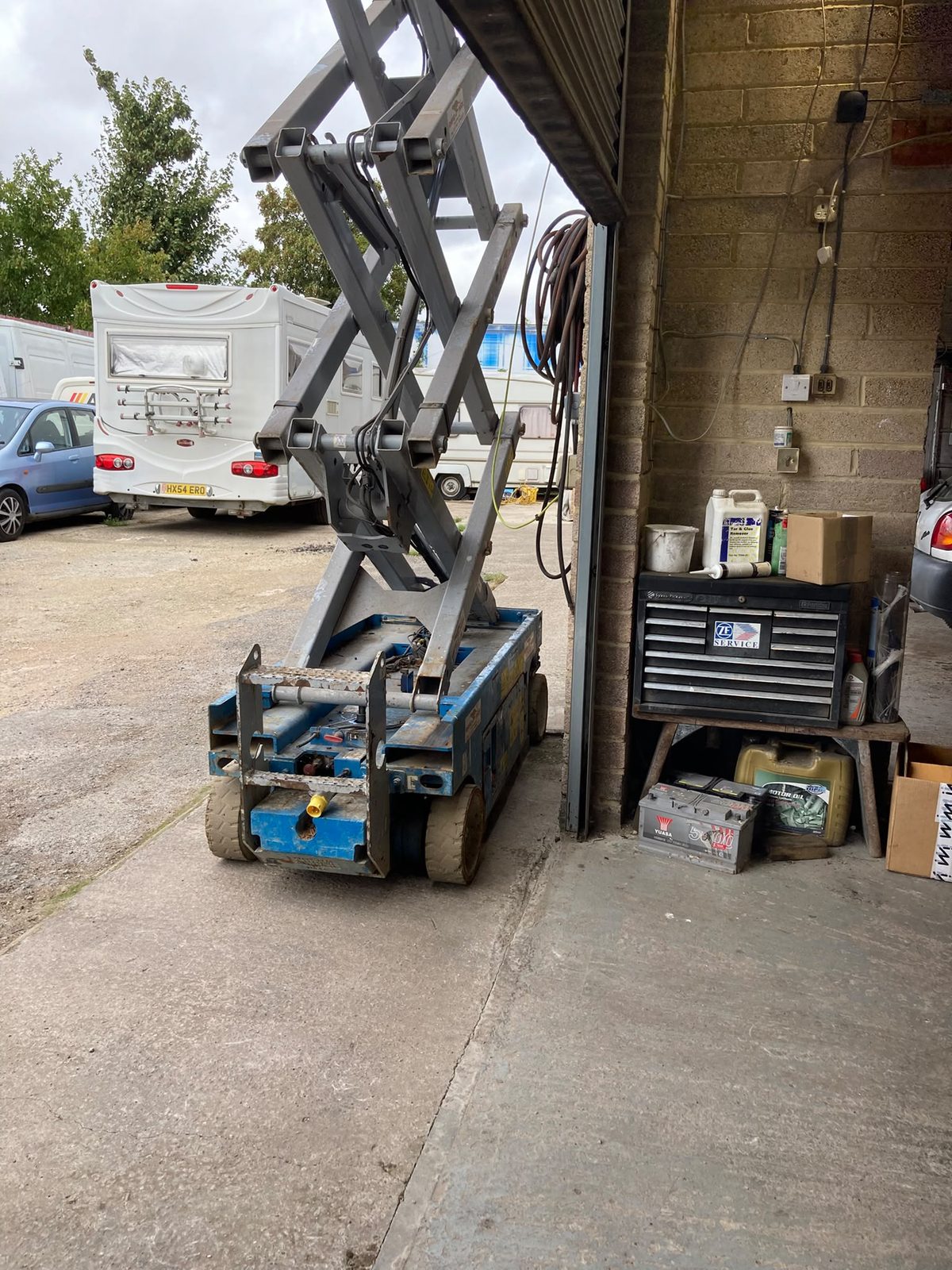 A blue scissor lift parked by the garage entrance with motorhomes and a tool chest nearby.