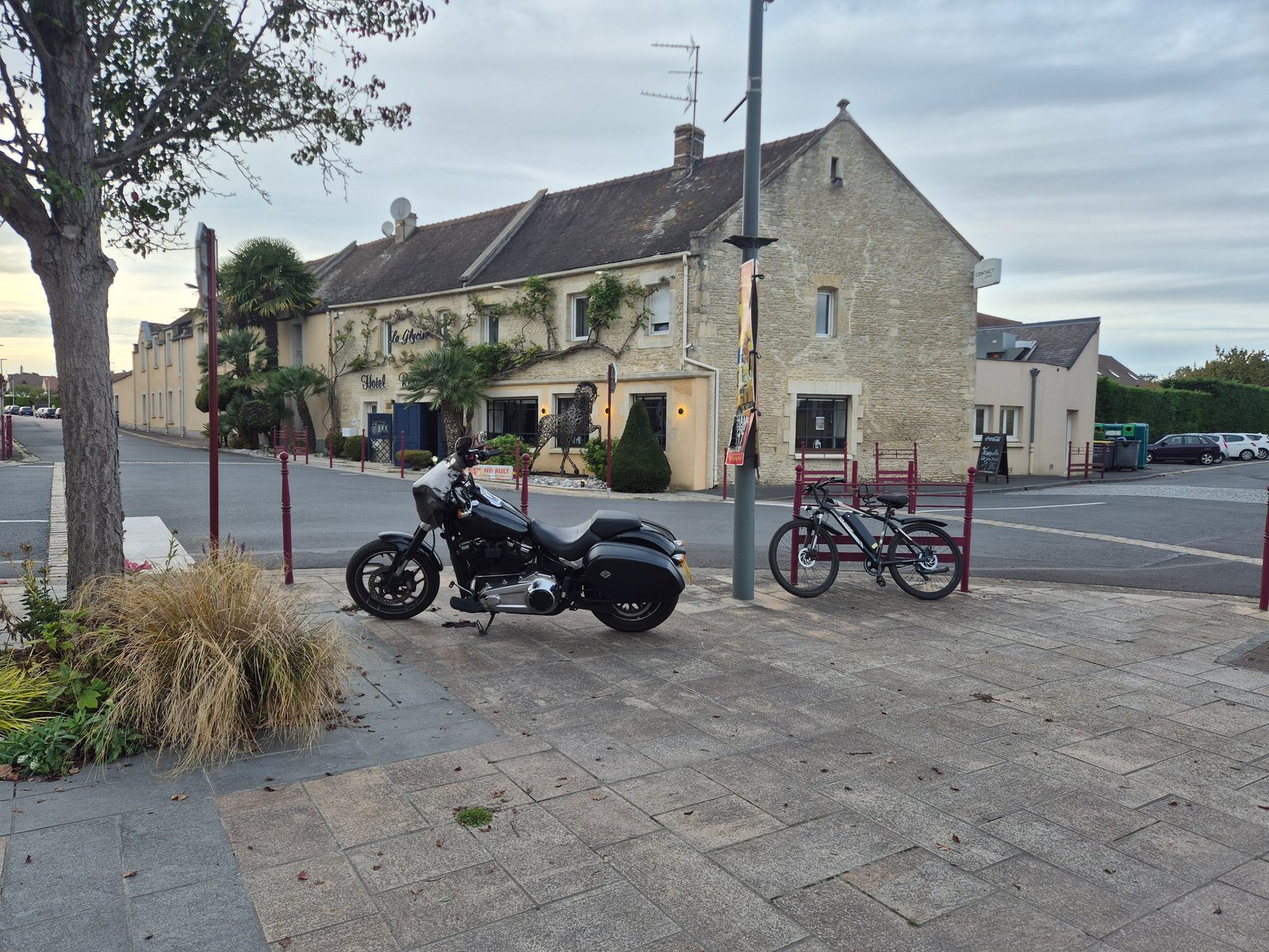 A black Harley-Davidson parked beside an electric bicycle on a paved square in front of a stone-built French hotel.