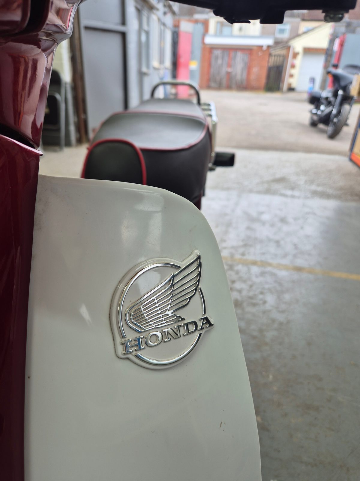 A chrome Honda winged logo on a cream bodywork panel, with a maroon scooter, mopeds and a motorcycle parked in a yard behind.
