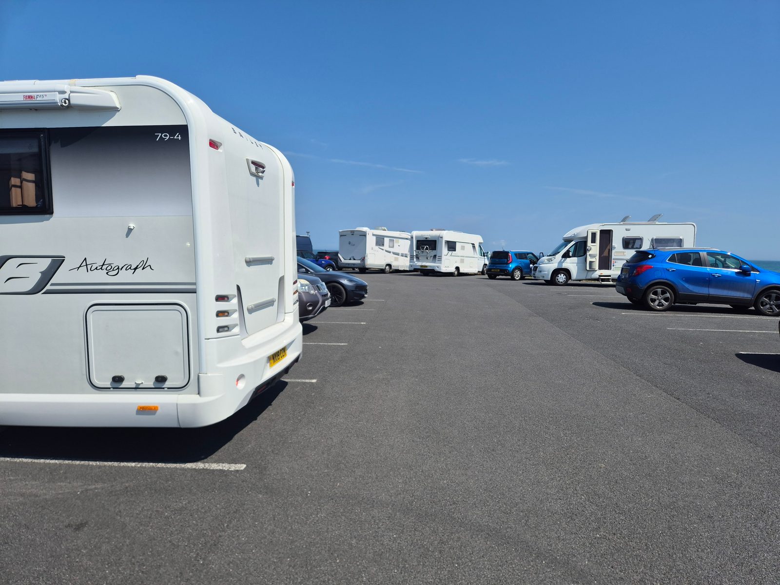 A white Bailey Autograph coachbuilt motorhome parked in a tarmac car park with other motorhomes and cars, under a clear blue sky by the sea.