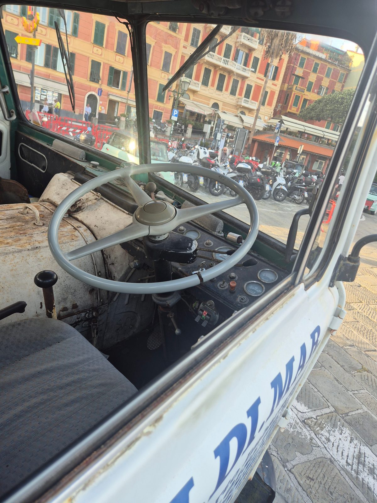 Interior of a vintage Italian work truck showing a large thin steering wheel and old dashboard, with a square of pastel buildings and parked scooters outside.