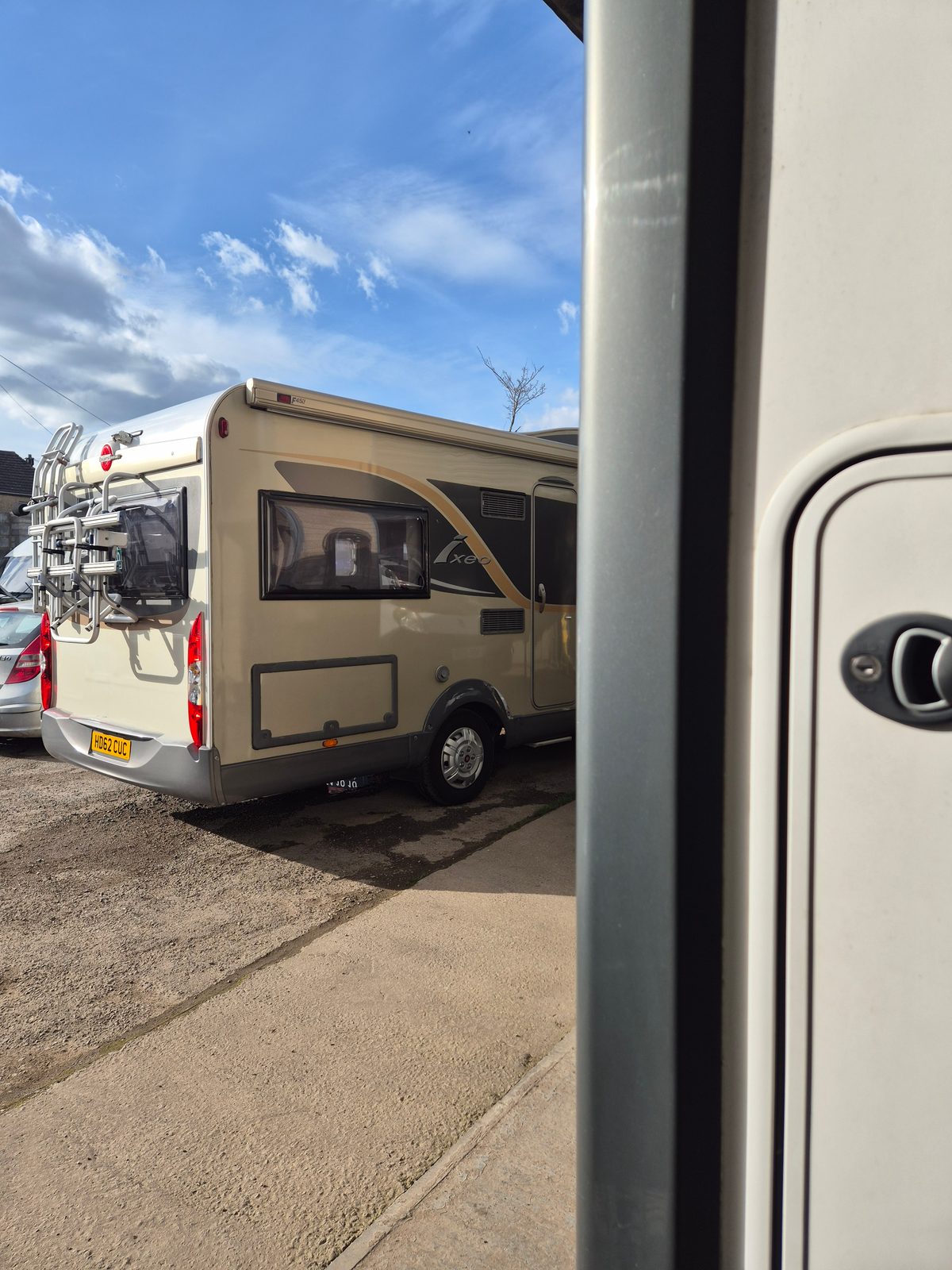 A rear view of a cream Burstner Ixeo coachbuilt motorhome with a rear bike rack, parked on gravel beside a silver car under a blue sky, with a motorhome bodywork panel and door in the foreground.