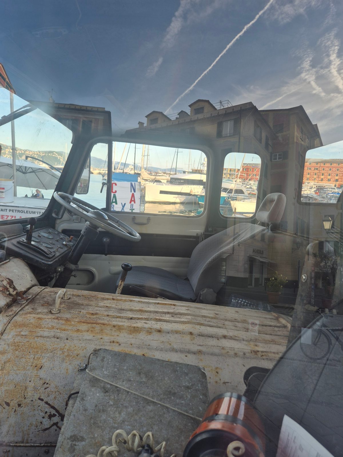 View through the window into the worn cab of an old truck, with a steering wheel and seat, overlooking a harbour full of boats and old buildings.
