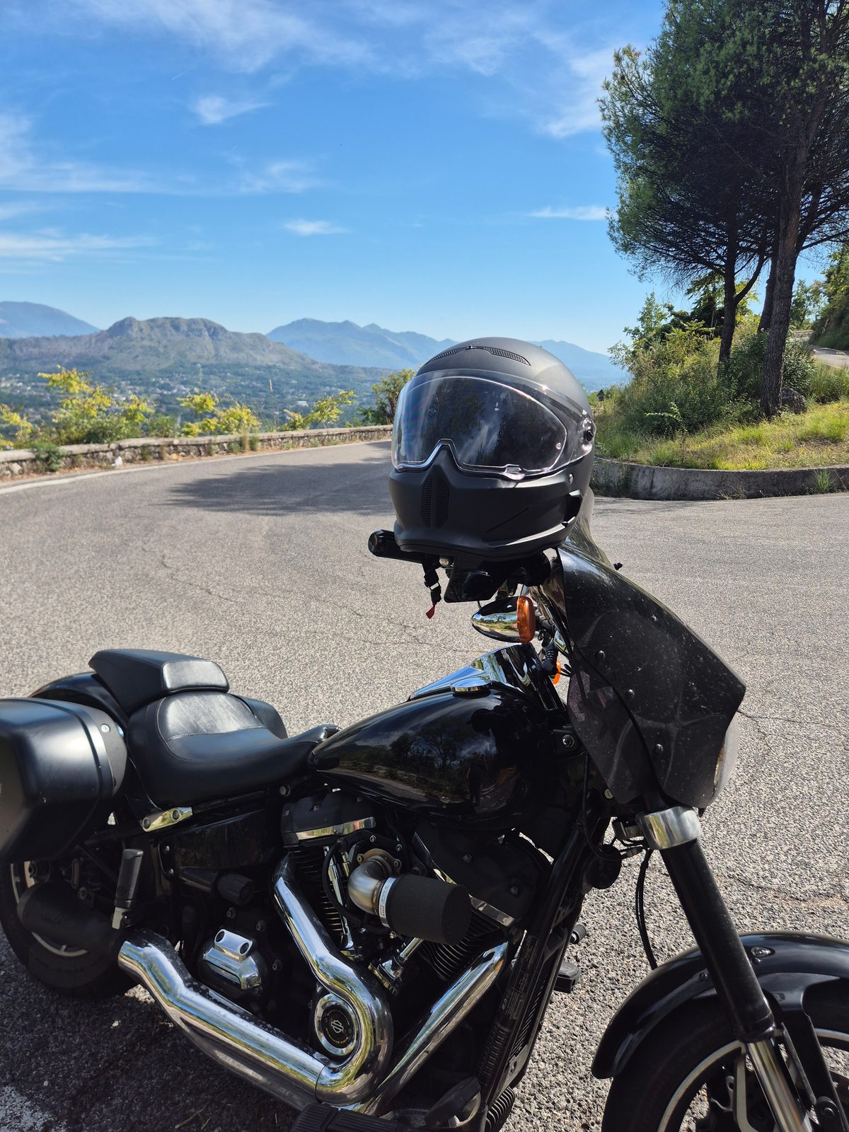 A black Harley-Davidson with a helmet resting on the handlebars parked on a mountain road with hills in the distance.