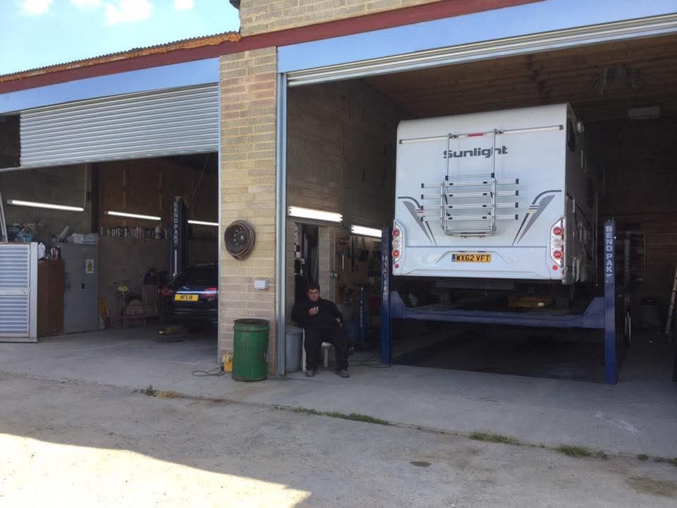 A white Sunlight motorhome raised on a four-post lift in an open garage bay, with a man seated outside and a black car on a ramp in the adjacent bay.