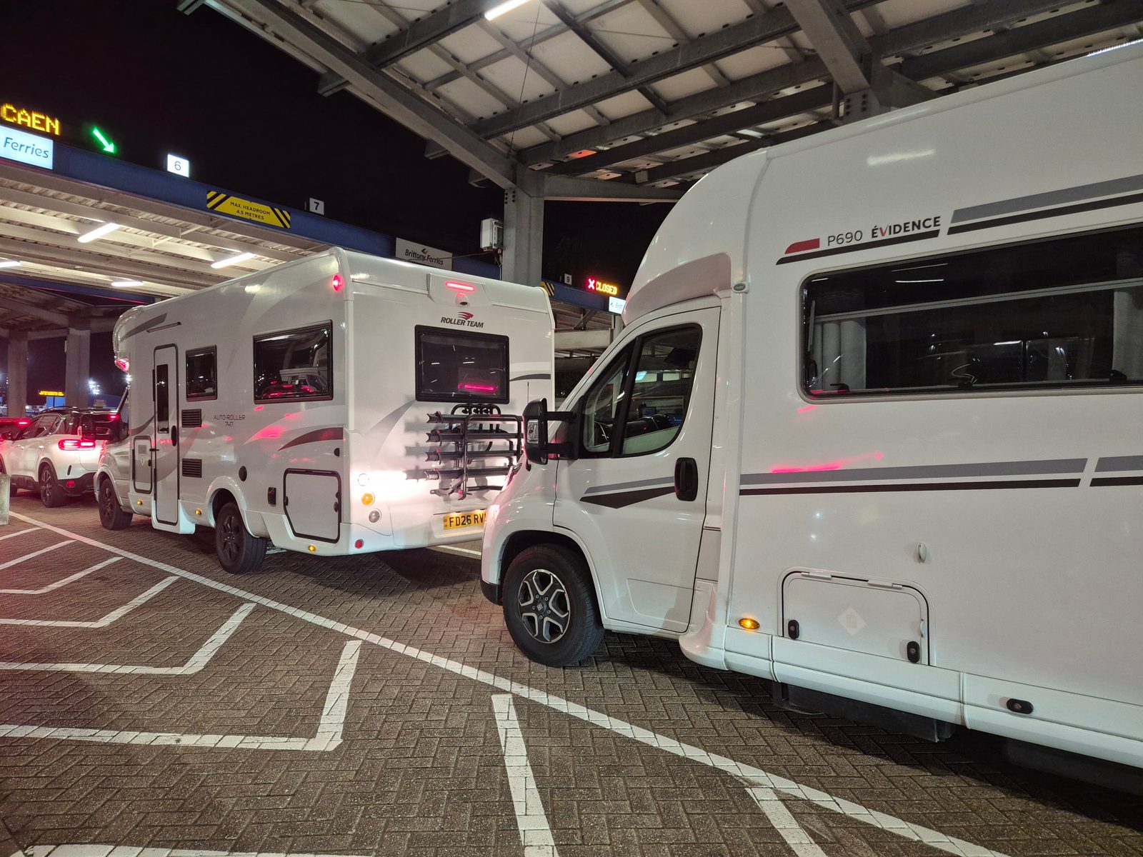 A white Roller Team Auto-Roller motorhome with a rear bike rack and a white P690 Evidence motorhome queuing under a covered ferry terminal at night, with a sign for Caen Brittany Ferries overhead.