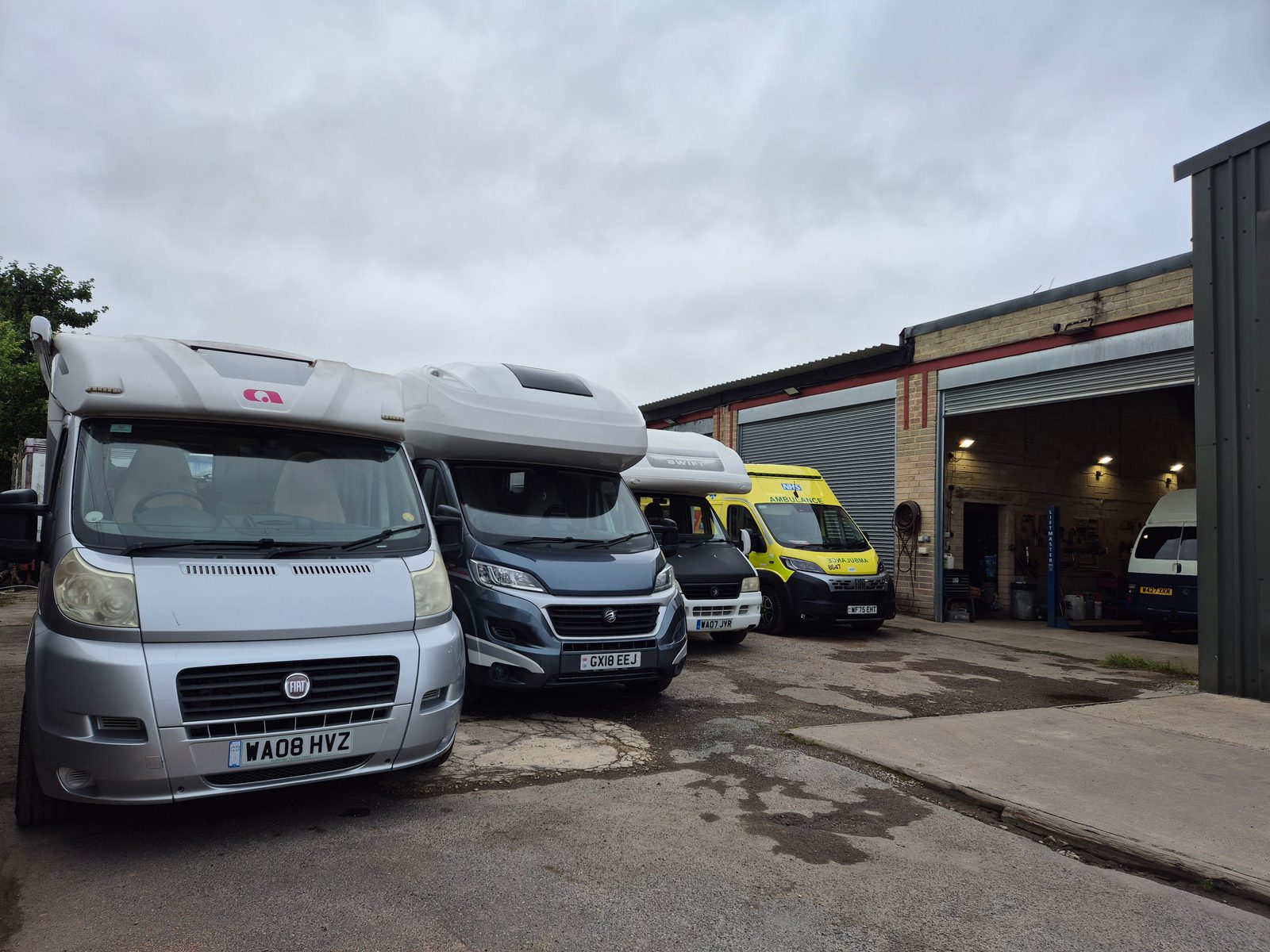 A row of coachbuilt Fiat motorhomes and a yellow NHS ambulance parked on the forecourt beside an open garage workshop with a van inside on a ramp.