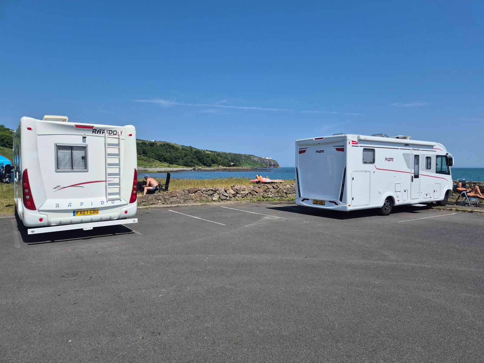 A white Rapido coachbuilt motorhome and a white Pilote motorhome parked in a coastal car park overlooking the sea, with people relaxing by a stone wall in the sunshine.