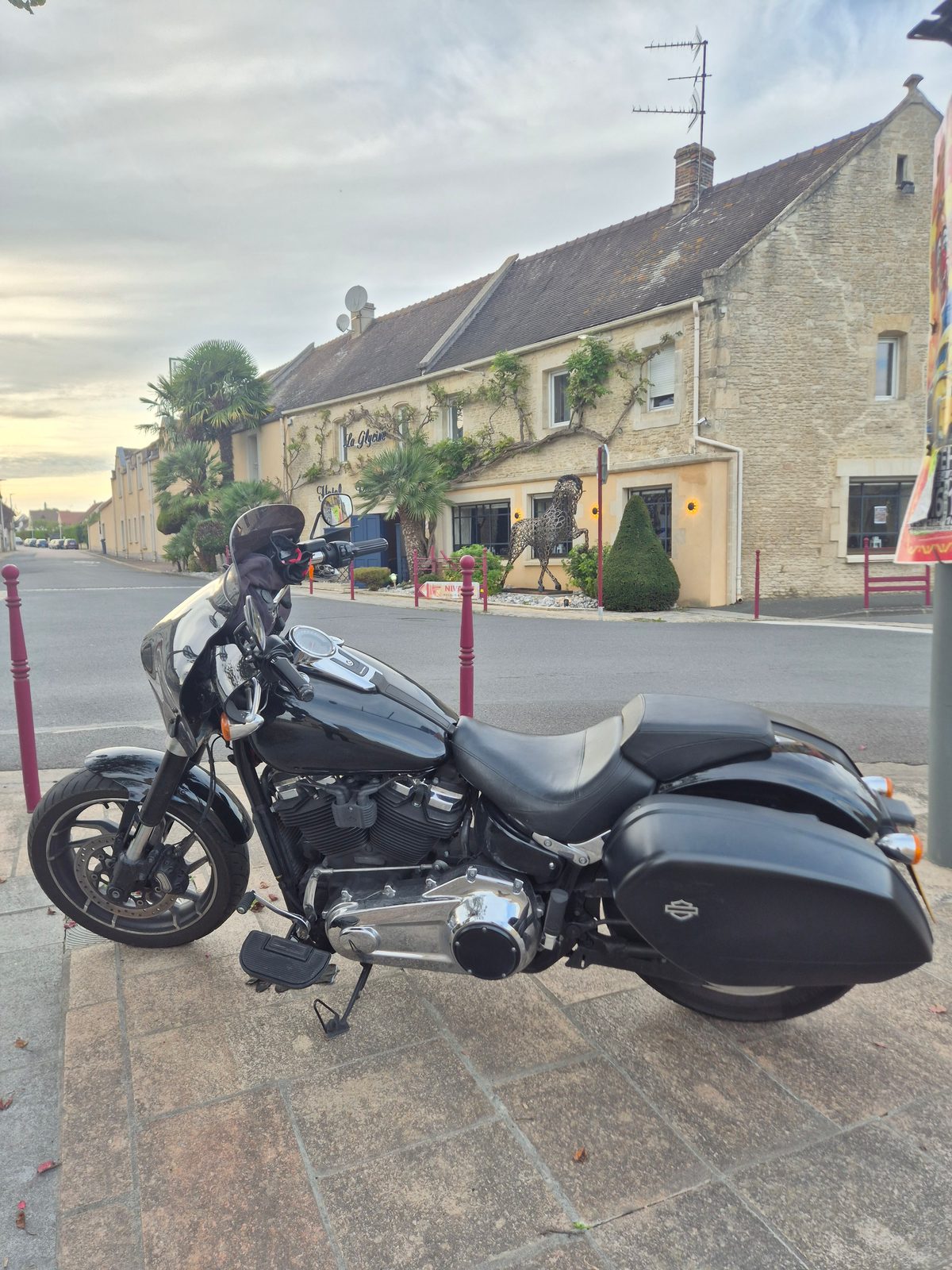 A black Harley-Davidson motorcycle with panniers parked on a pavement outside a stone hotel in a French town at dusk.