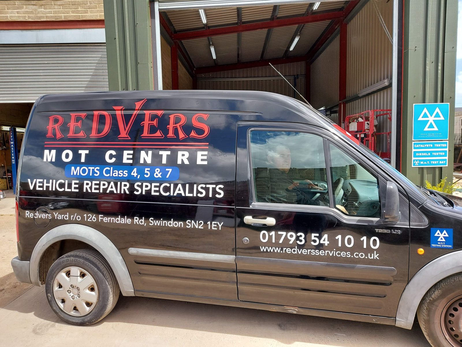 Black Ford Transit Connect van with Redvers MOT Centre signwriting parked outside the garage.