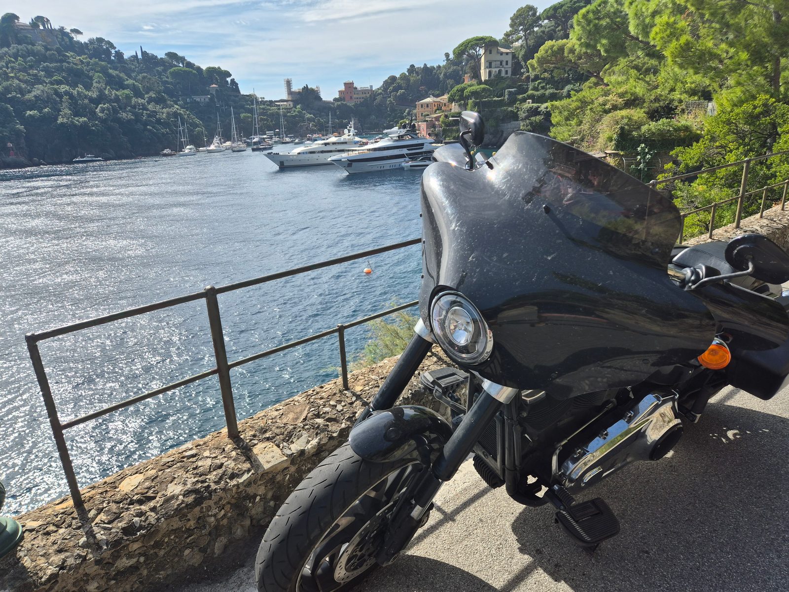 A black motorcycle parked by a railing above a harbour full of yachts and motor boats, with wooded hills and villas beyond.