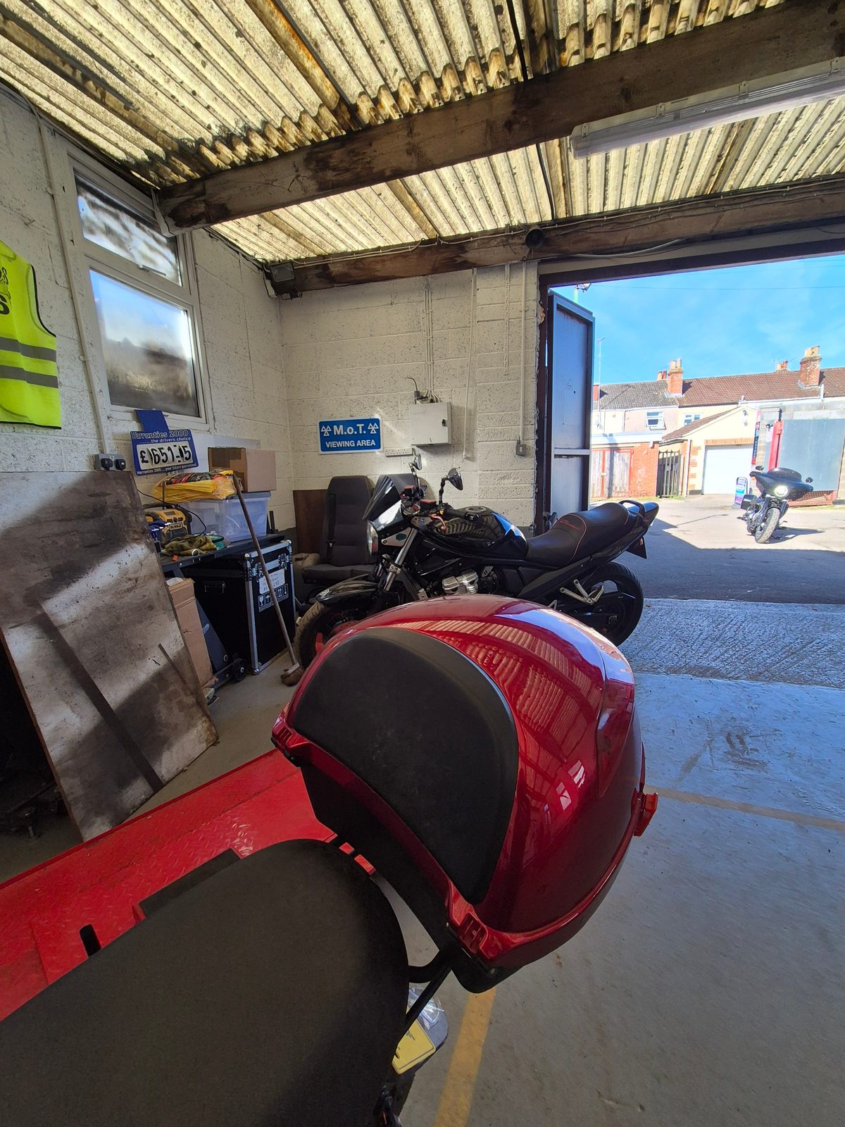 Several motorcycles queued inside a garage for MOT testing, with the roller door open to a sunny street.