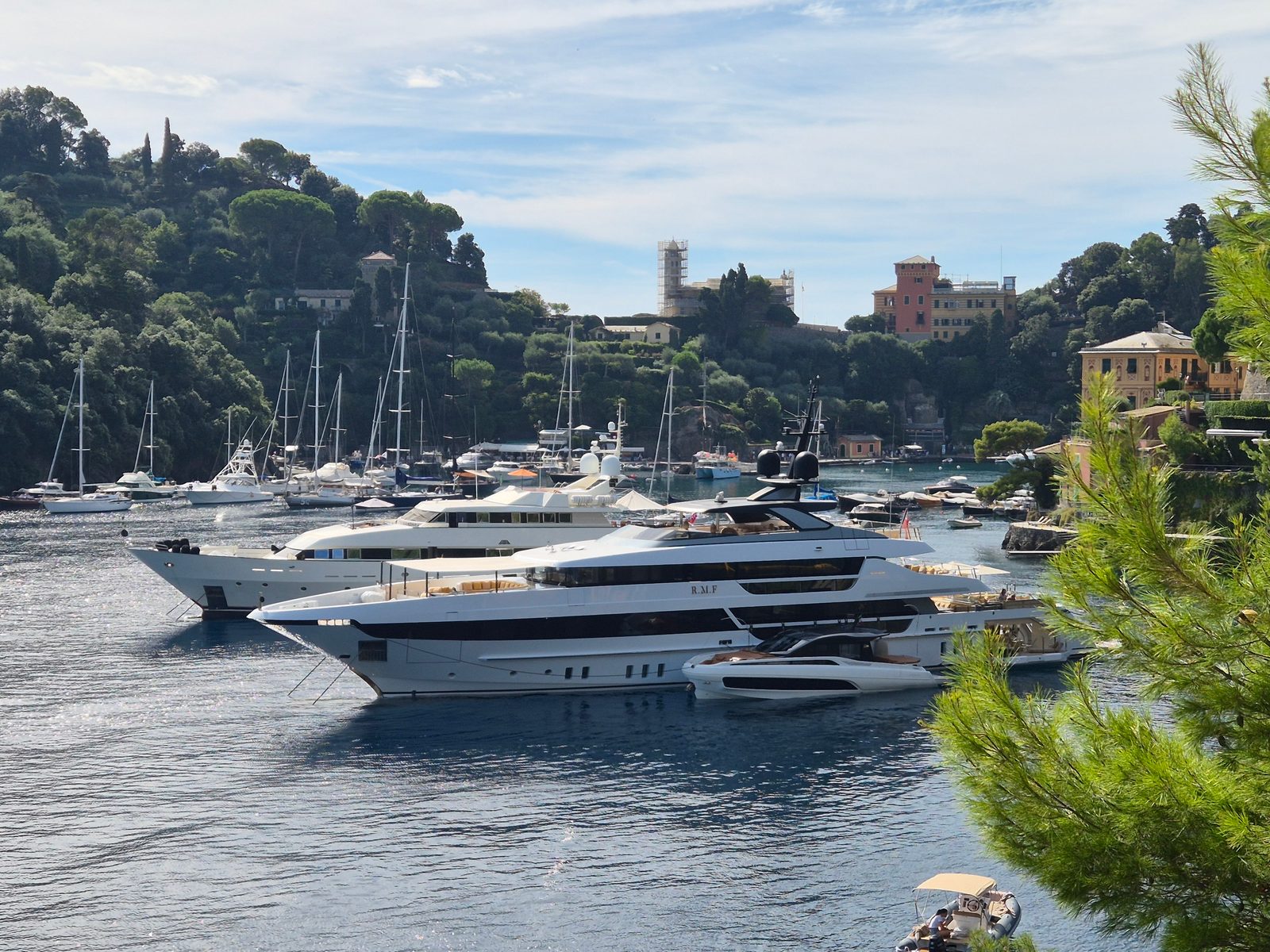 Large white superyachts moored in a sheltered Italian harbour with wooded hills and colourful villas behind.