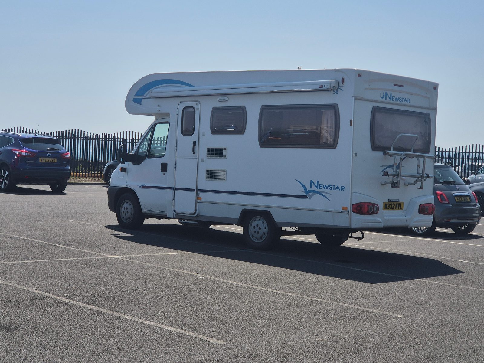 A white Fiat-based Newstar coachbuilt motorhome with an overcab bed and rear bike rack, parked in a tarmac car park beside a blue Nissan and a Fiat 500, with metal railings behind.