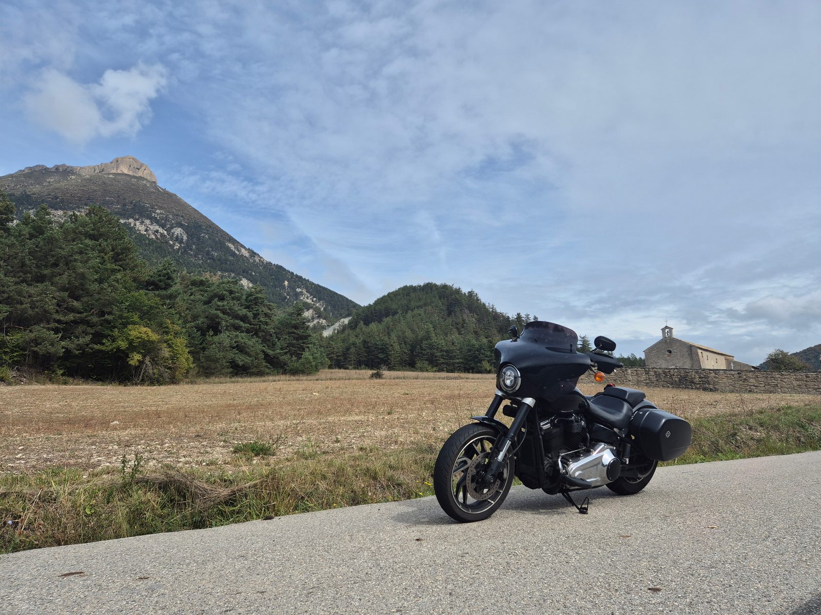 A black Harley-Davidson parked on a quiet mountain road beside a field, with a stone chapel and rocky peaks behind.