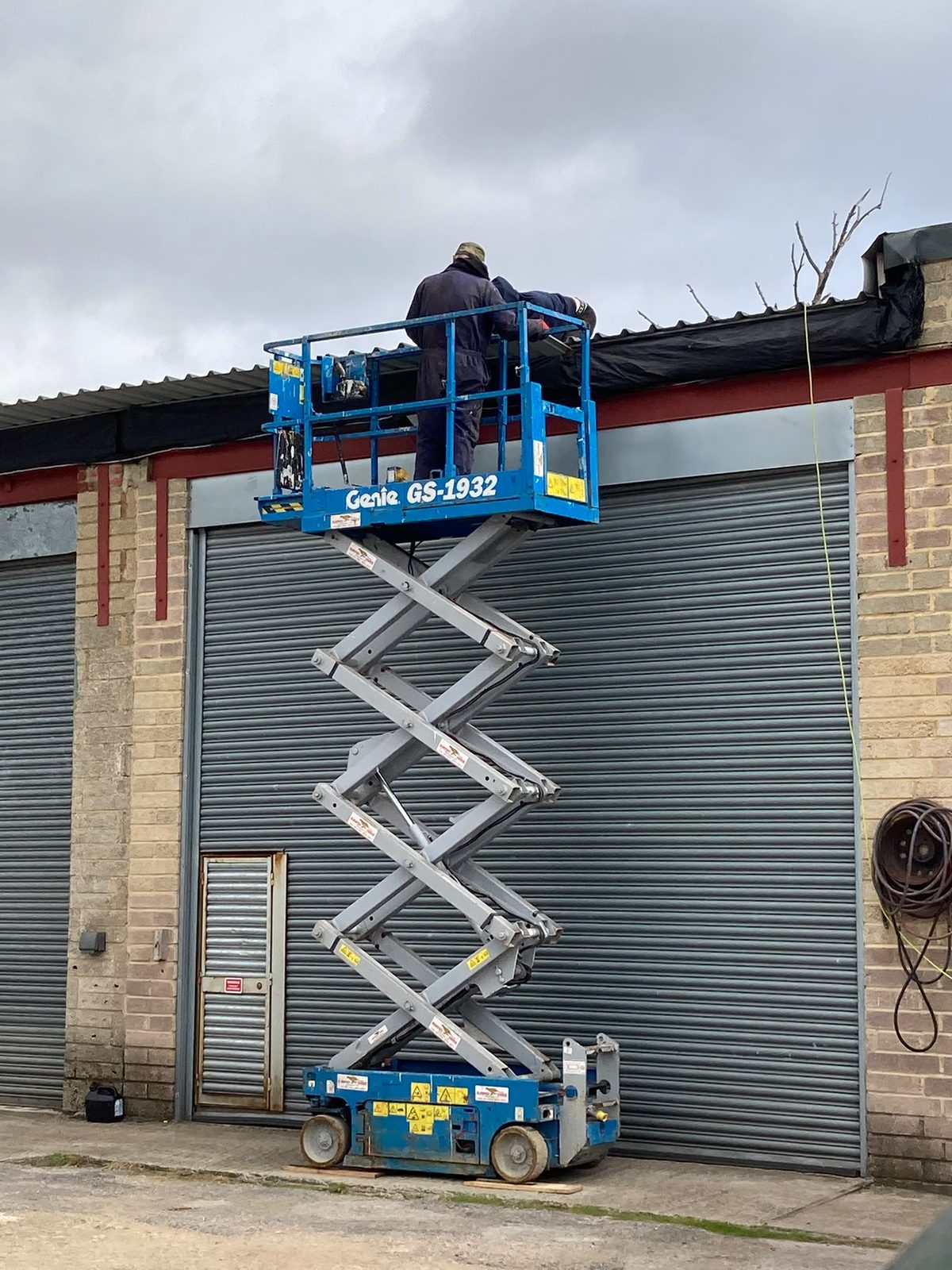 Two men working on a roof from a raised blue Genie scissor lift outside the garage.