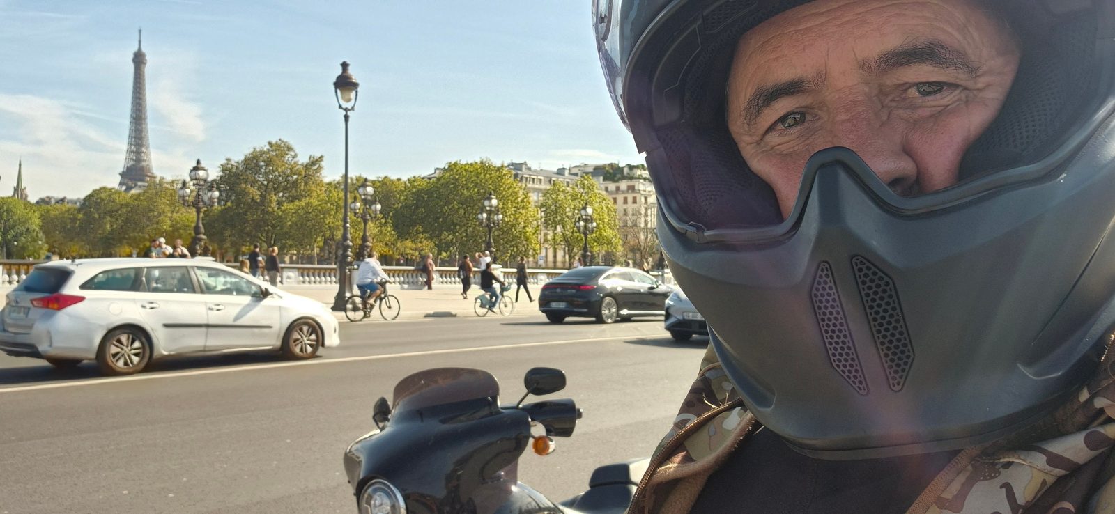 A man wearing a motorcycle helmet with face mask on his bike, with the Eiffel Tower and Paris traffic behind him.