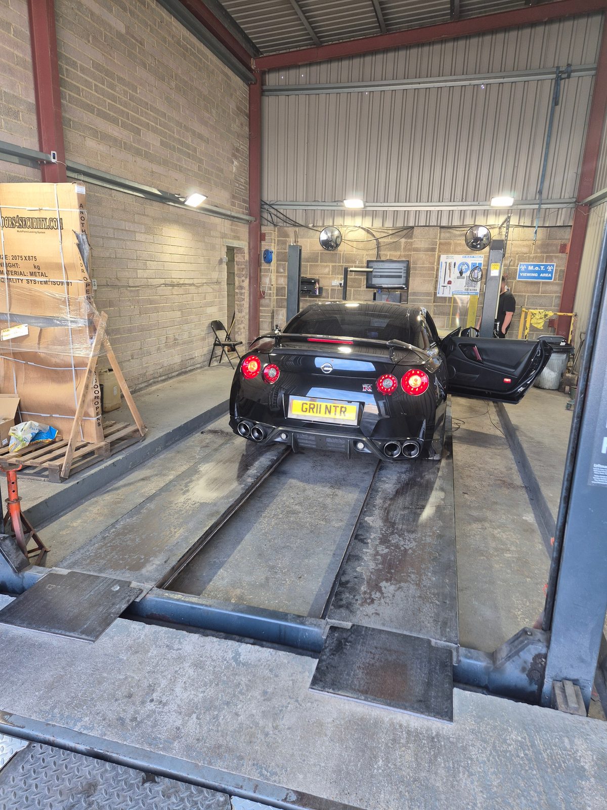 A black Nissan GT-R coupe with driver's door open, parked over an MOT inspection bay with a viewing area sign on the brick wall behind.