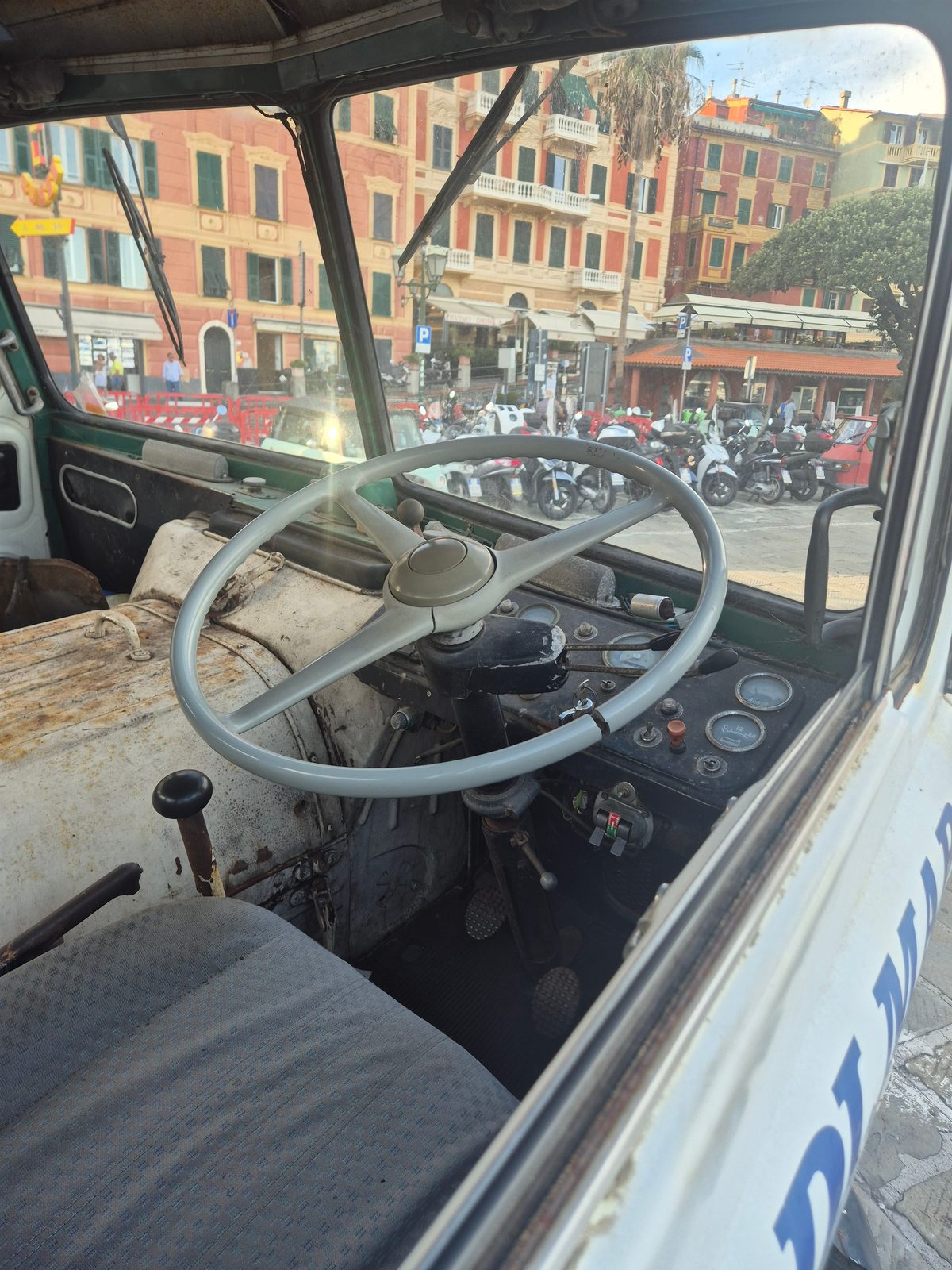 View from inside the cab of a vintage commercial vehicle with a large grey steering wheel, looking out onto an Italian town square with parked scooters and colourful buildings.