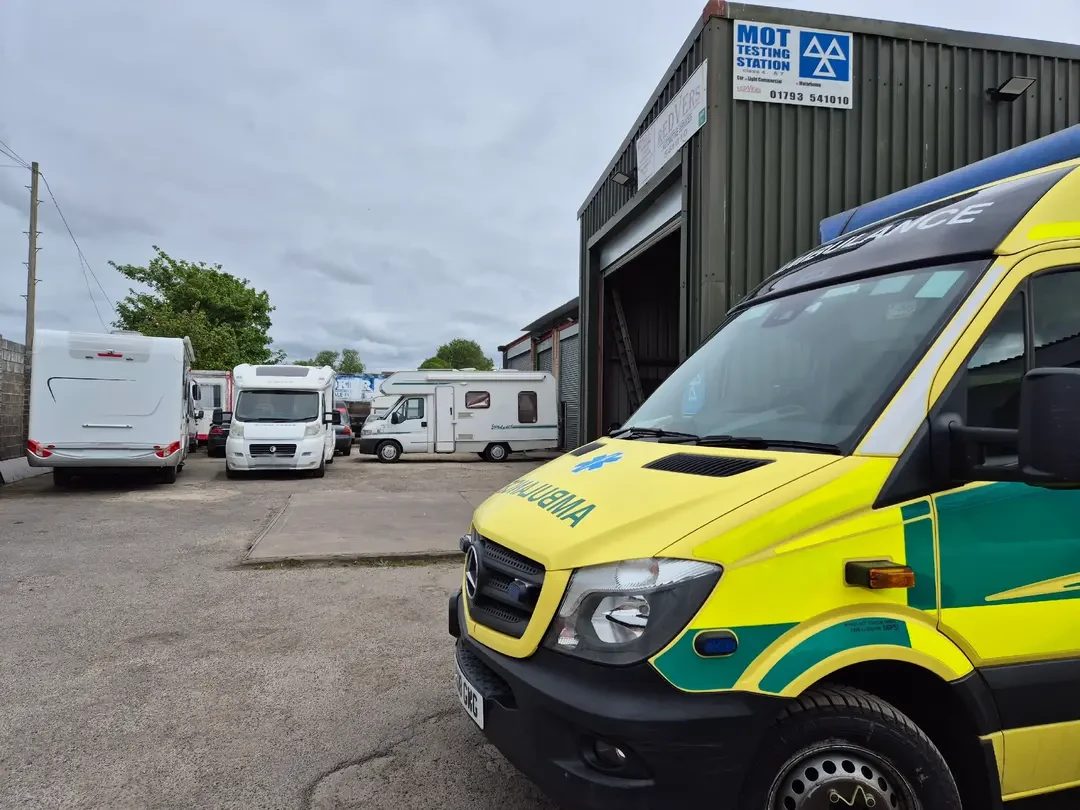 A Mercedes-Benz Sprinter ambulance in yellow and green livery parked beside a grey industrial unit with an MOT Testing Station sign, with several motorhomes lined up to the left.