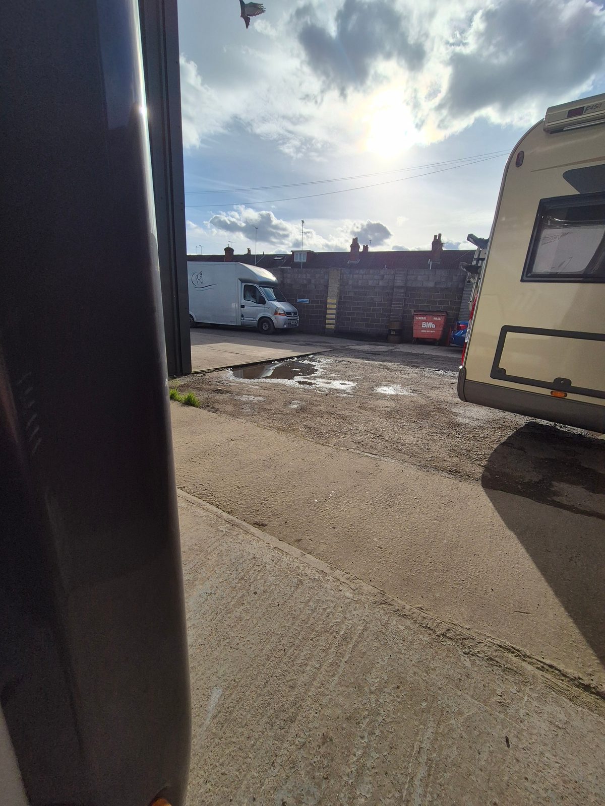 A caravan parked to the right and a white box van beyond a brick wall, seen from a garage doorway over a concrete yard with puddles and a Biffa waste bin.