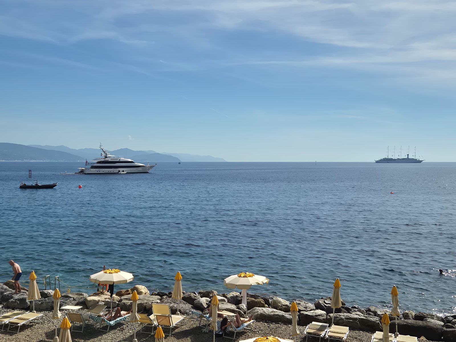 A rocky beach lined with yellow sunloungers and parasols looking out over a calm blue sea with yachts anchored offshore and hills in the distance.