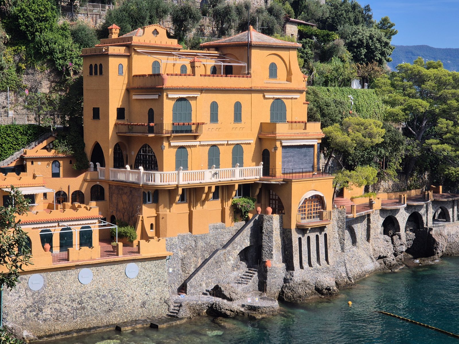 A large orange Italian villa built into the rocks above clear turquoise sea, with arched windows and green shutters.