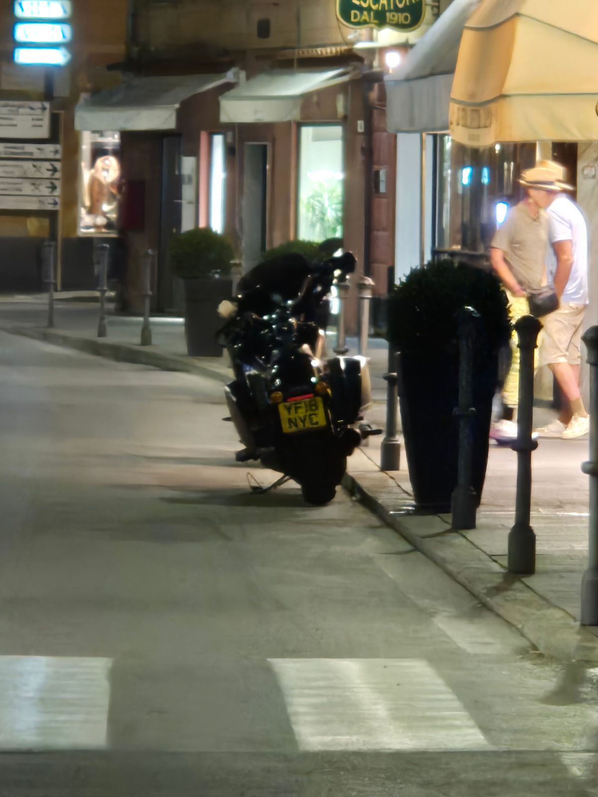 A black motorcycle with a UK number plate parked on a street at night outside shops with a couple walking past.