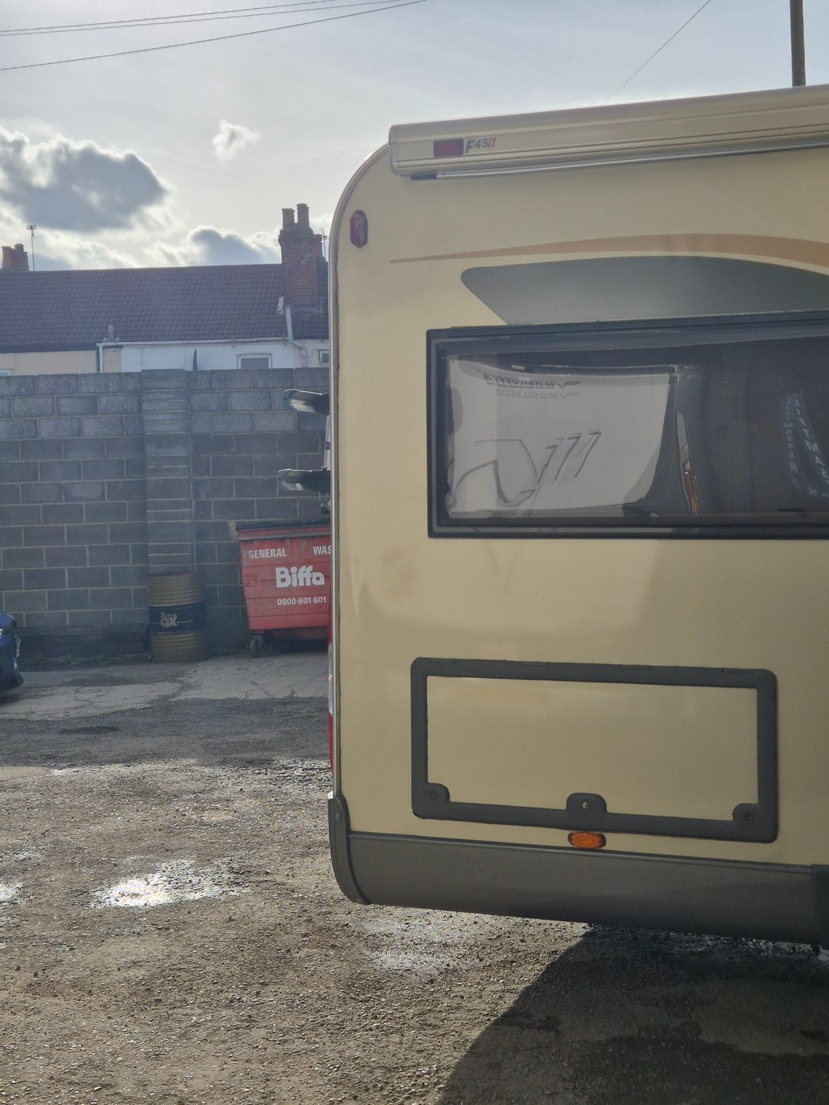 The rear corner of a cream coachbuilt motorhome with a Fiamma awning and window, parked on a gravel yard beside a breeze-block wall and a red Biffa skip.
