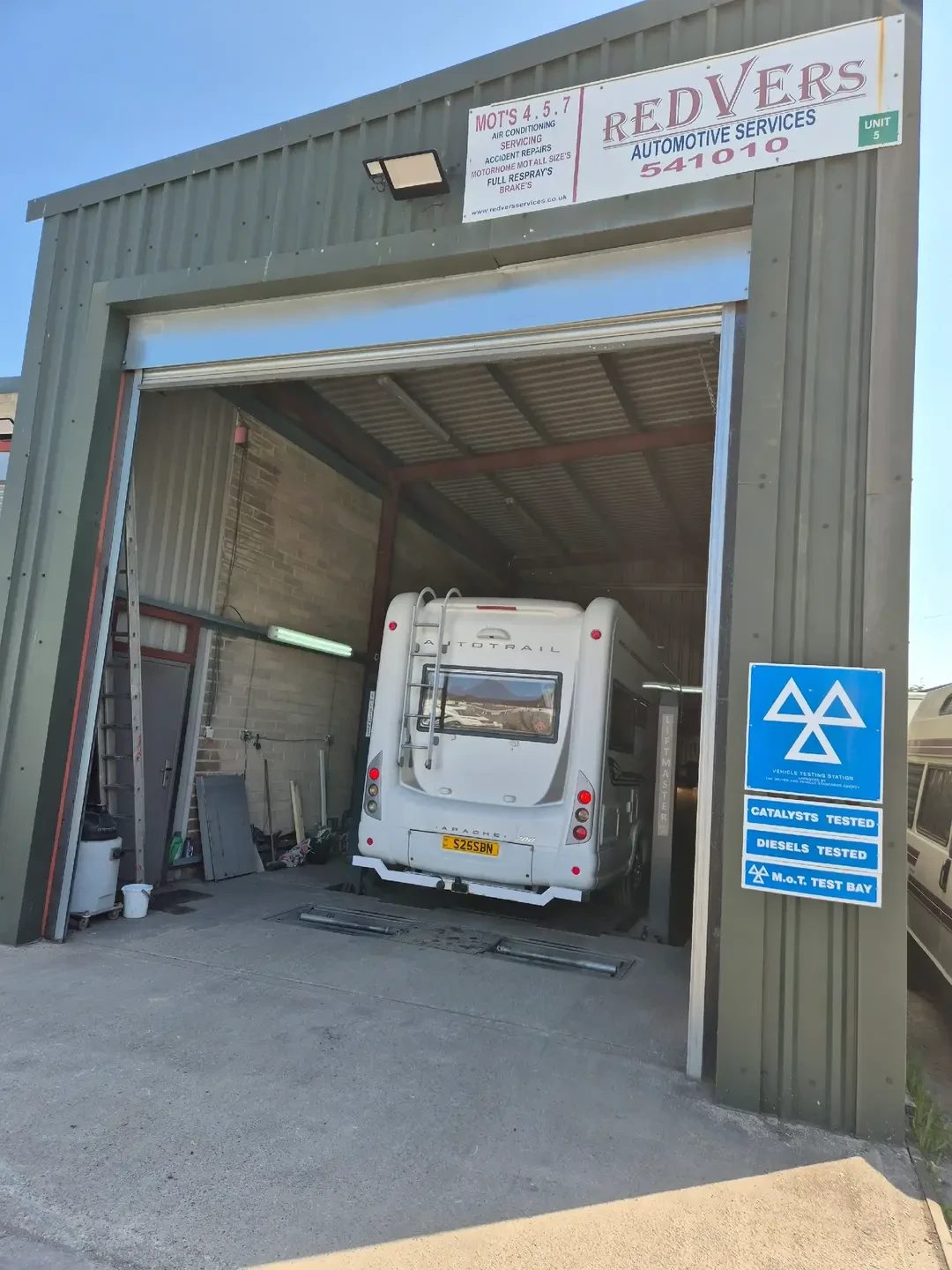 A white Auto-Trail Apache coachbuilt motorhome parked in the Redvers Automotive Services workshop, with signage reading Redvers Automotive Services and MOT test bay.