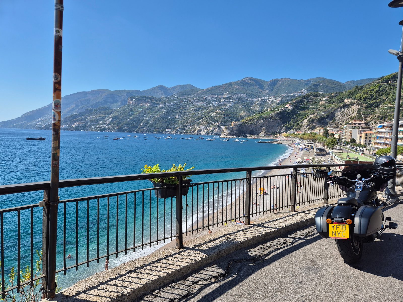 A motorcycle parked by railings overlooking a bay with boats, a beach and mountains behind.