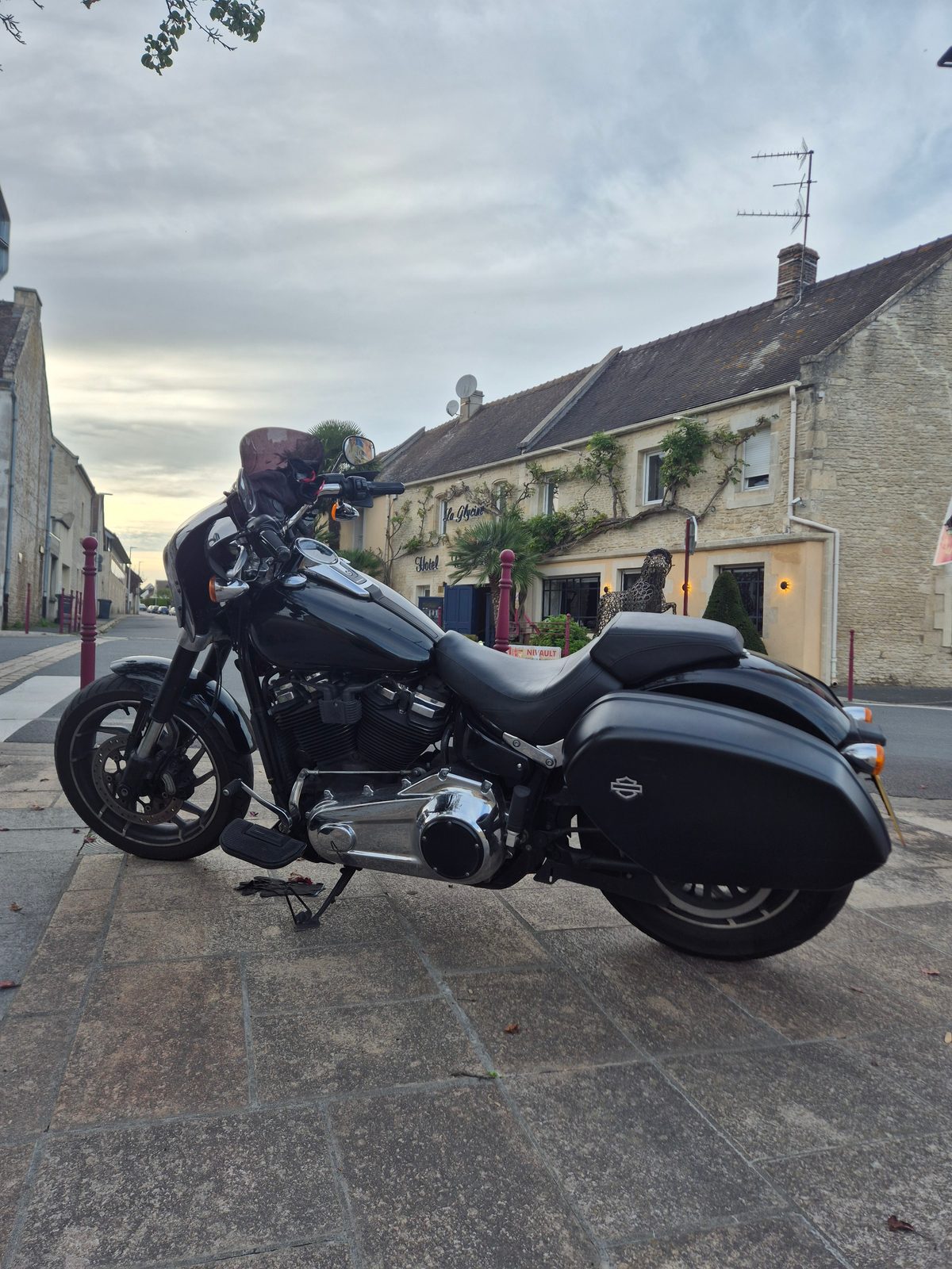 A black Harley-Davidson motorcycle parked on a stone pavement outside a French stone hotel at dusk.