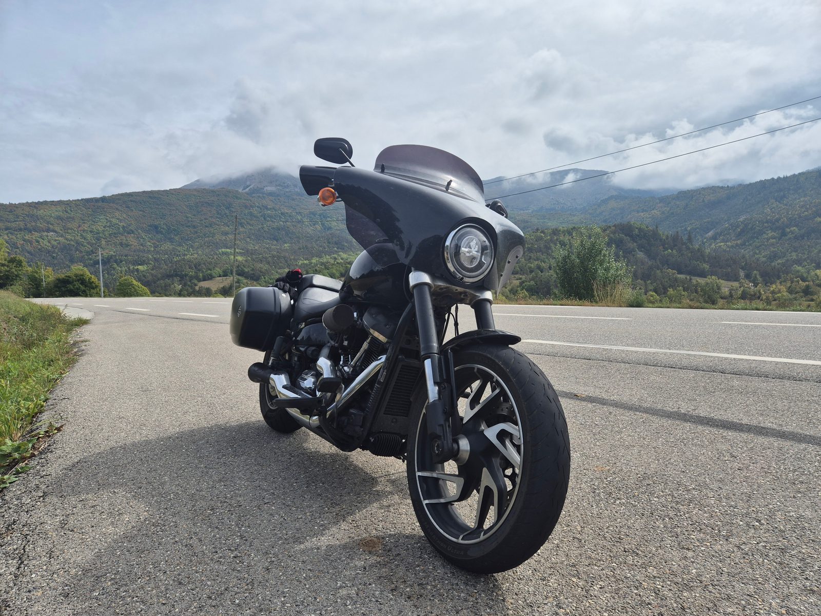 A black Harley-Davidson with fairing and panniers parked on a mountain road with cloud-covered peaks behind.