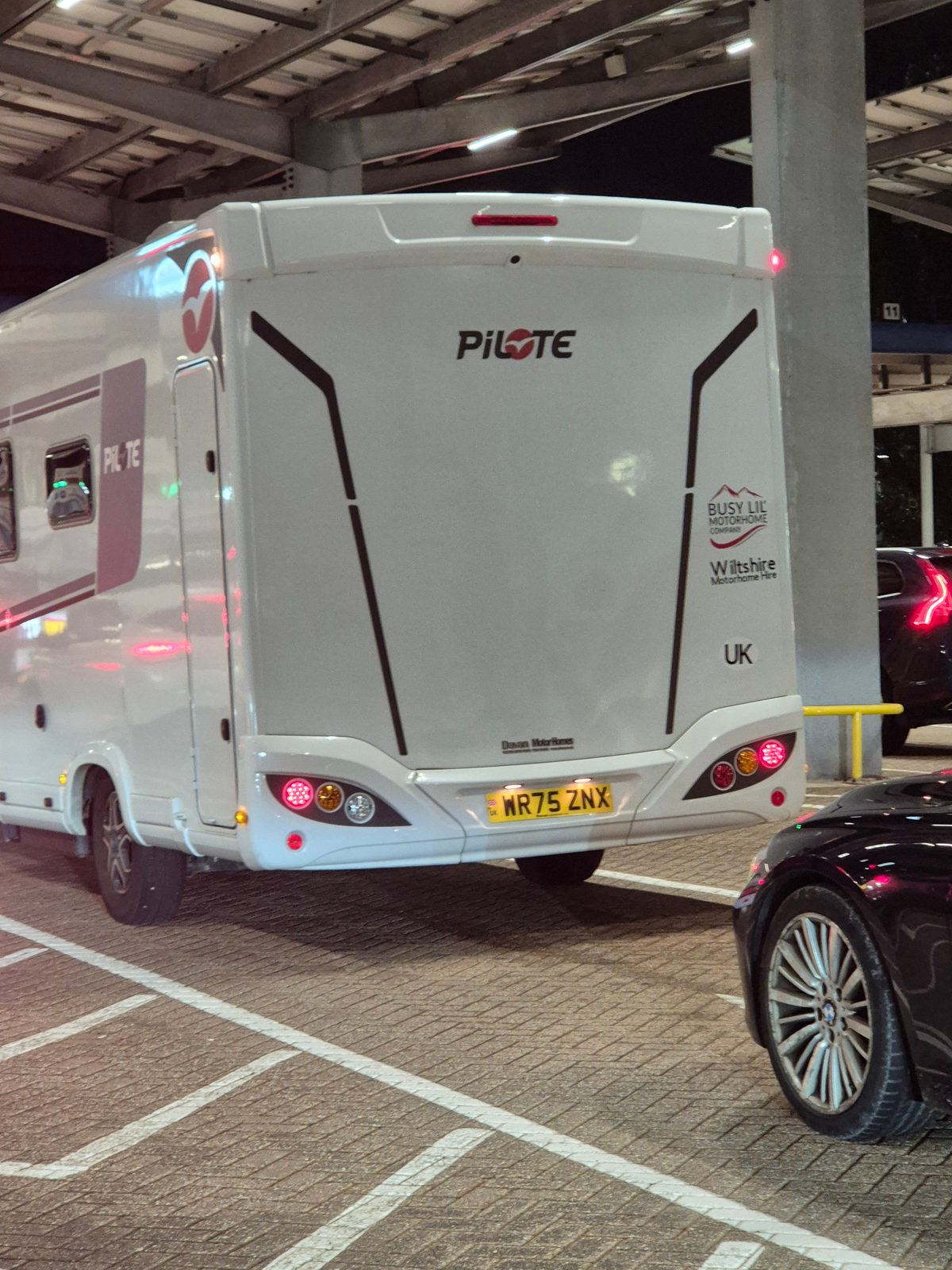 The rear of a white Pilote coachbuilt motorhome registered WR75 ZNX, parked in a covered multi-storey car park beside a black car at night.