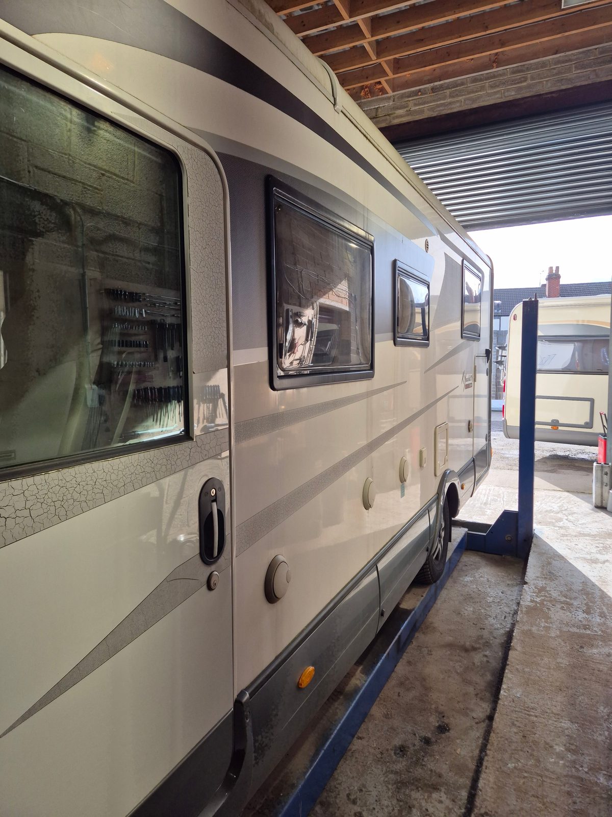 A white and grey coachbuilt motorhome parked on a ramp inside the garage, with a caravan visible through the open roller door.