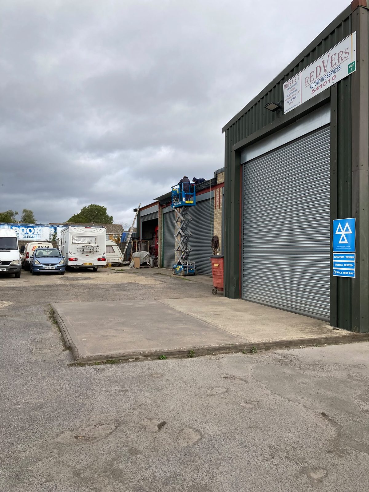Two workers on a blue scissor lift working on the roof of the Redvers garage unit, with vans and a motorhome parked in the yard under a cloudy sky.