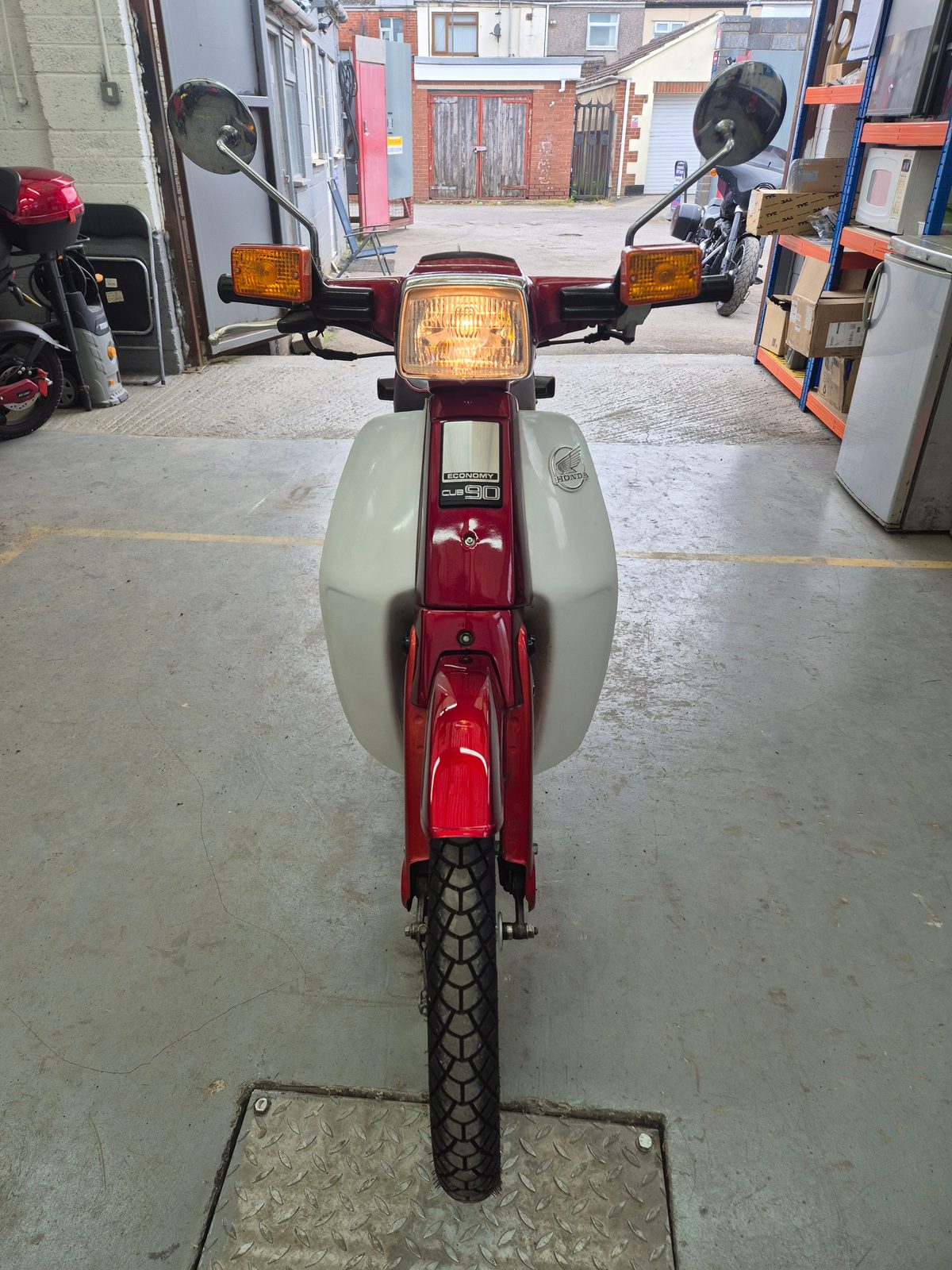 A red Honda Economy Cub 90 moped seen head-on with its headlamp on, parked over a metal floor plate inside a garage.