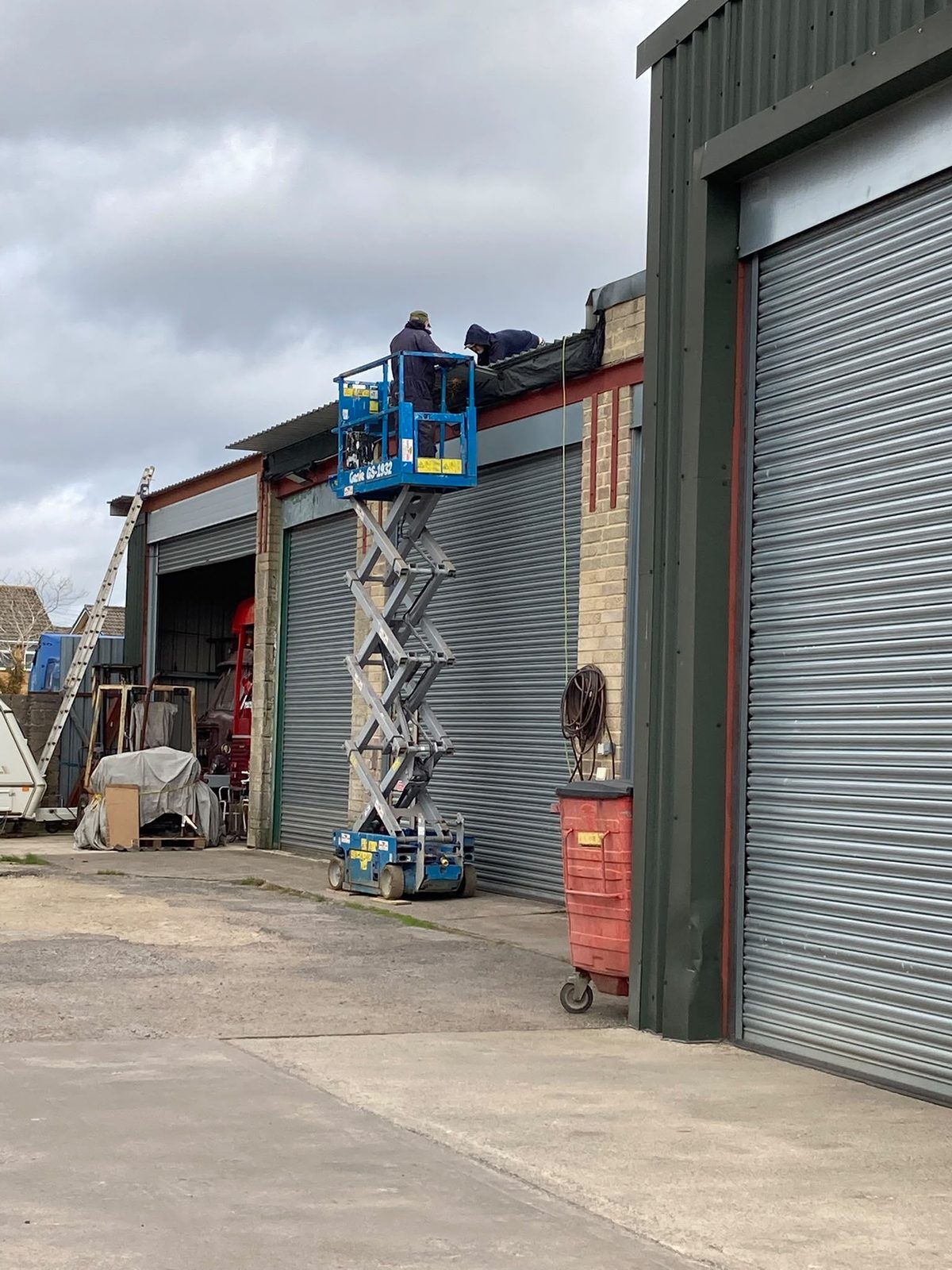 Two men on a raised blue scissor lift working on the roof edge of an industrial garage building with roller shutter doors.