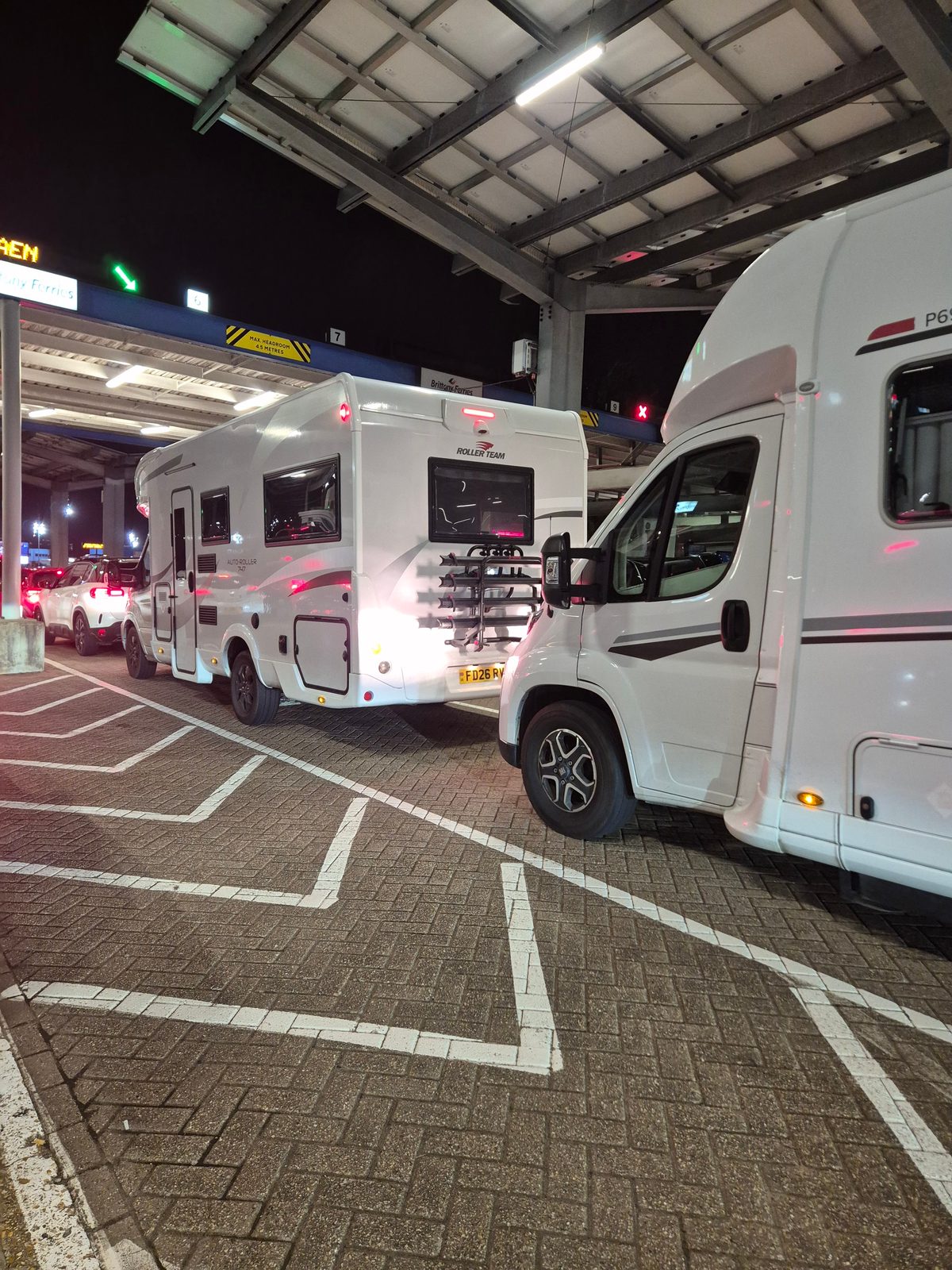 A white Roller Team coachbuilt motorhome with a rear bike rack queuing alongside another motorhome under a covered ferry terminal lane at night.