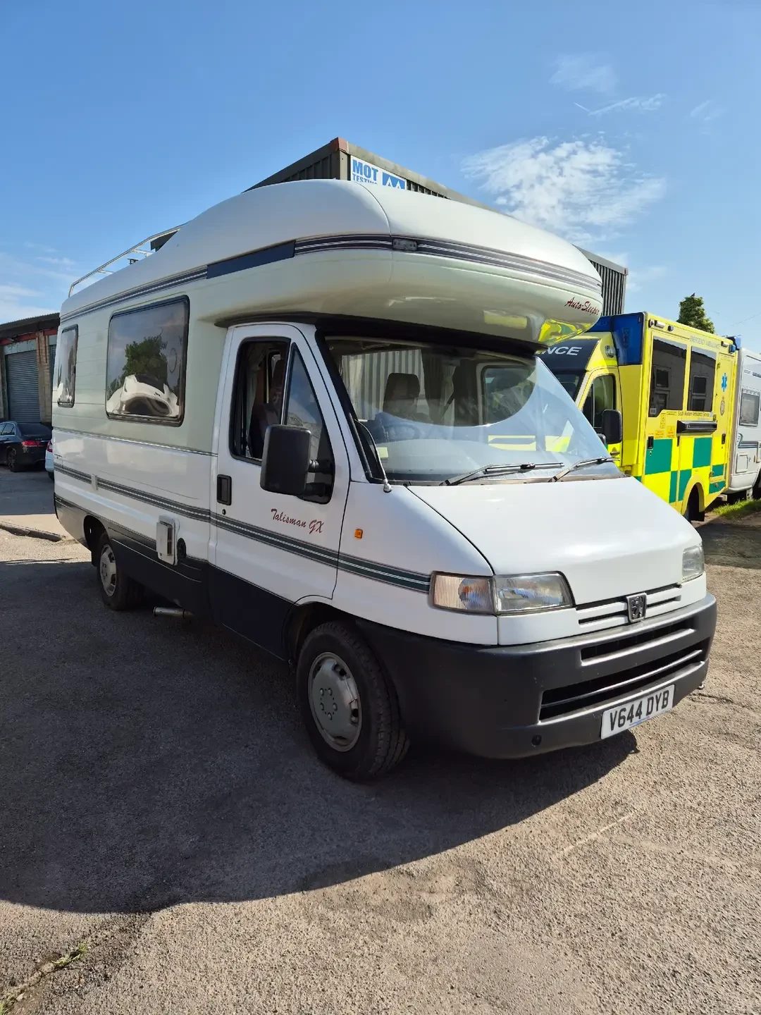 A white Peugeot-based Auto-Sleeper Talisman GX coachbuilt motorhome parked on gravel, with a yellow ambulance and MOT testing sign behind.