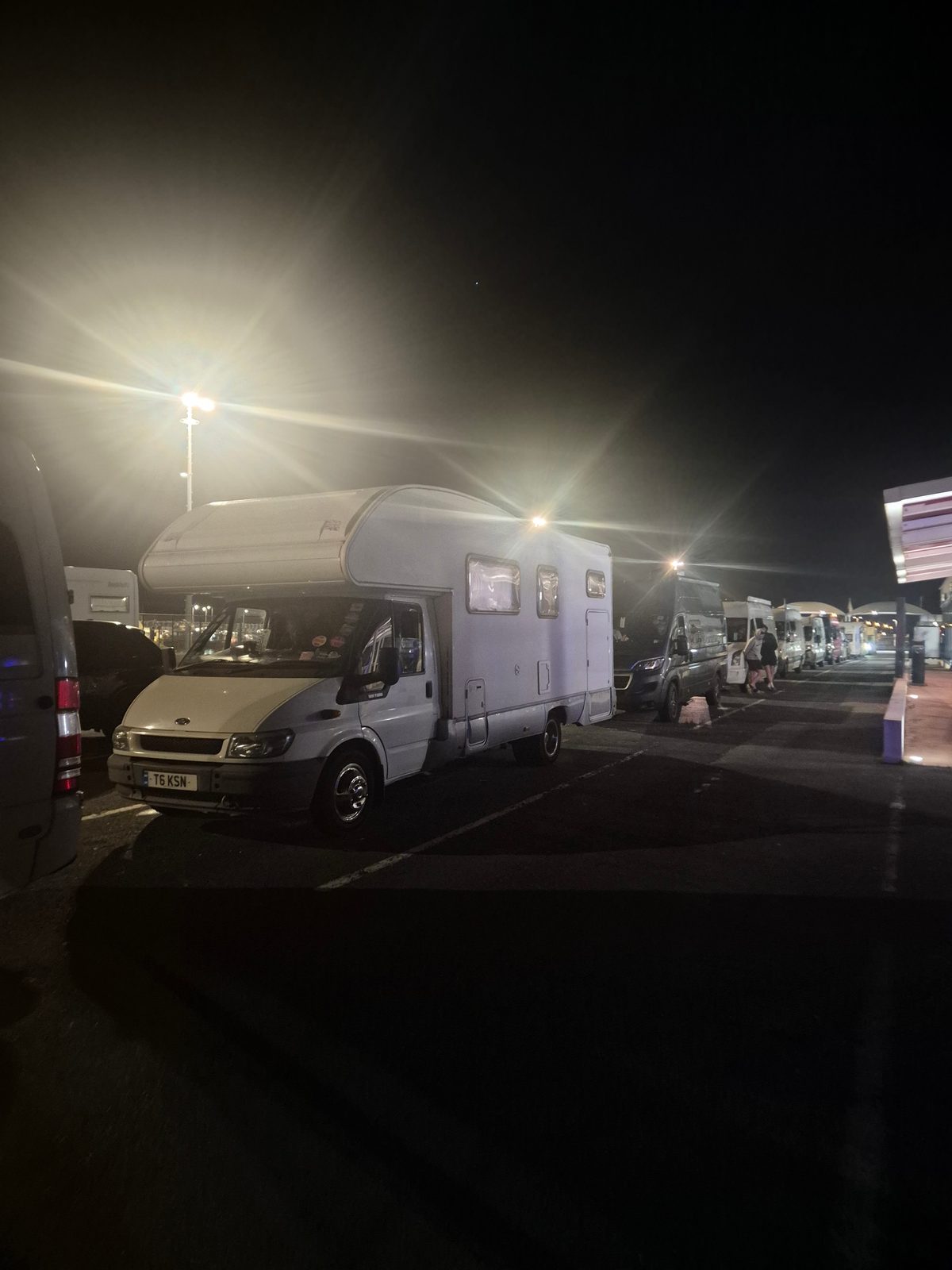 A white coachbuilt motorhome parked at night in a ferry terminal queue alongside other campervans under floodlights.