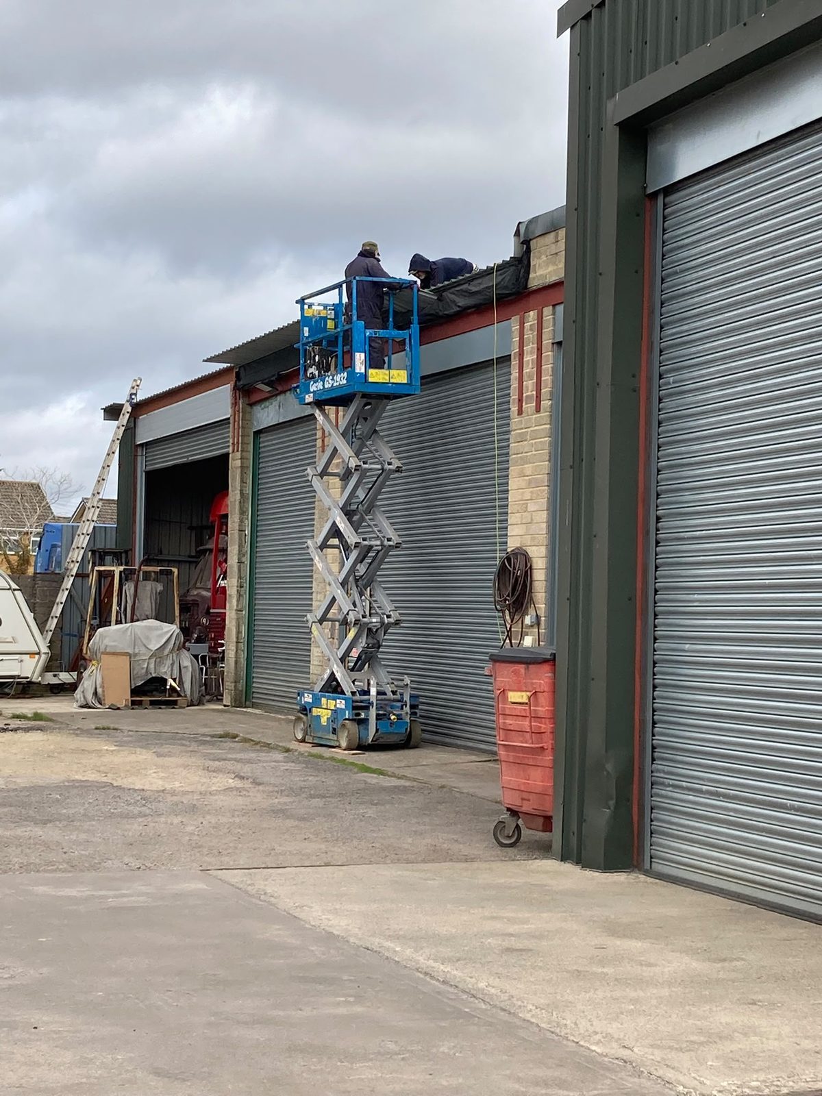 Two workers on a blue scissor lift repairing the roof above a garage shutter door.