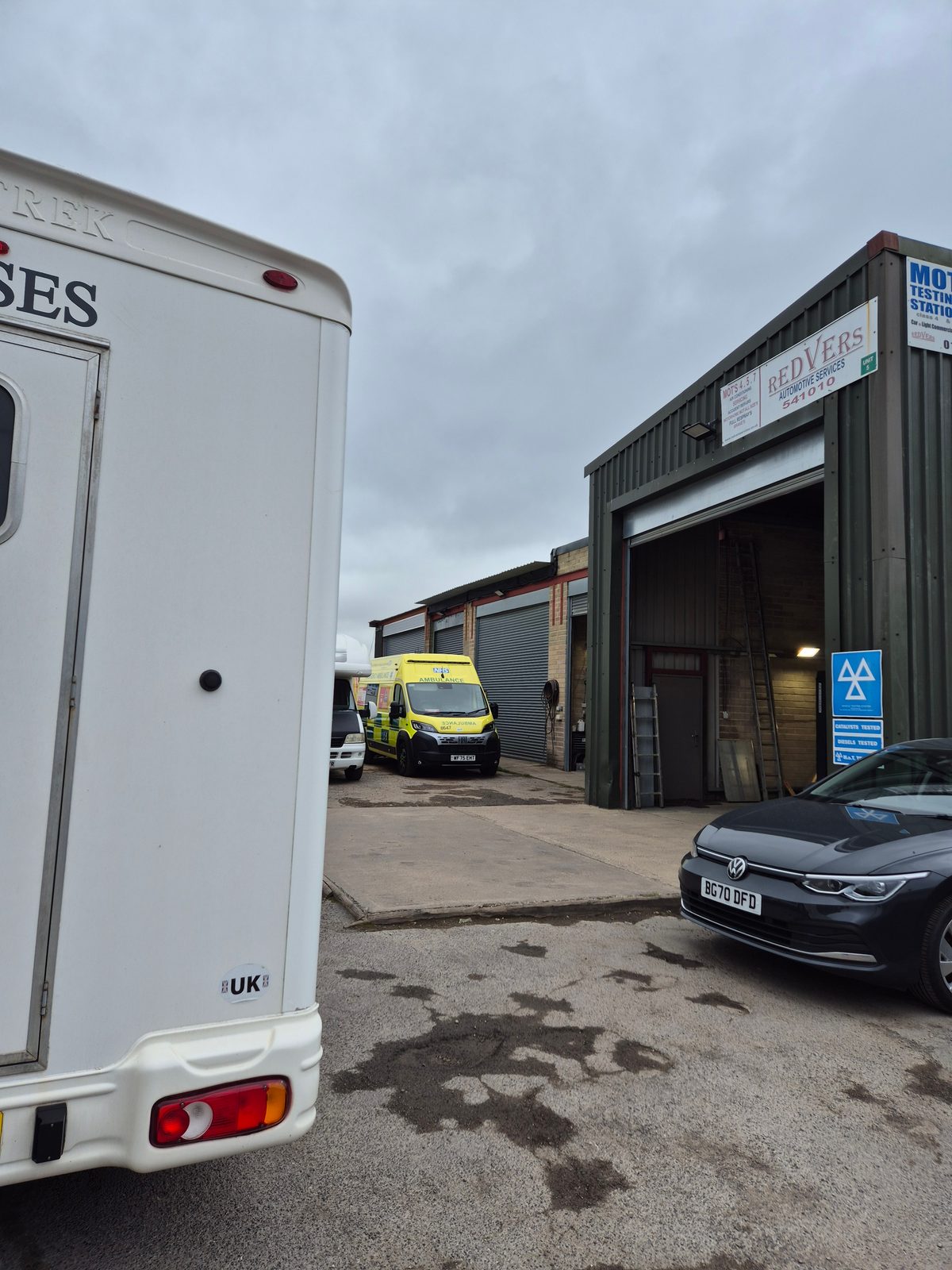 A white horsebox, a yellow NHS ambulance and a dark grey Volkswagen Golf parked outside a green industrial unit with Redvers Automotive Services and MOT Testing Station signage and an open workshop door.