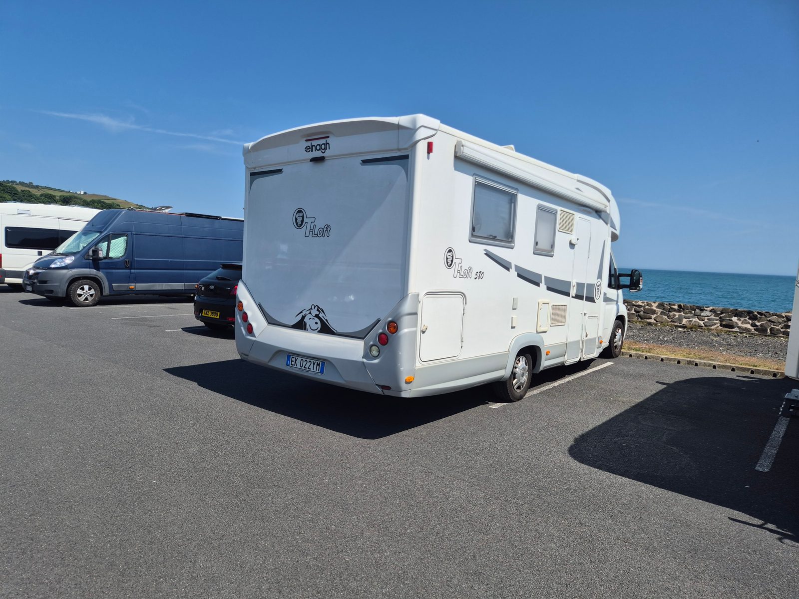 A white Elnagh T-Loft 530 coachbuilt motorhome with Italian plates parked in a seafront car park, with a dark van and a car nearby and the sea in the background.