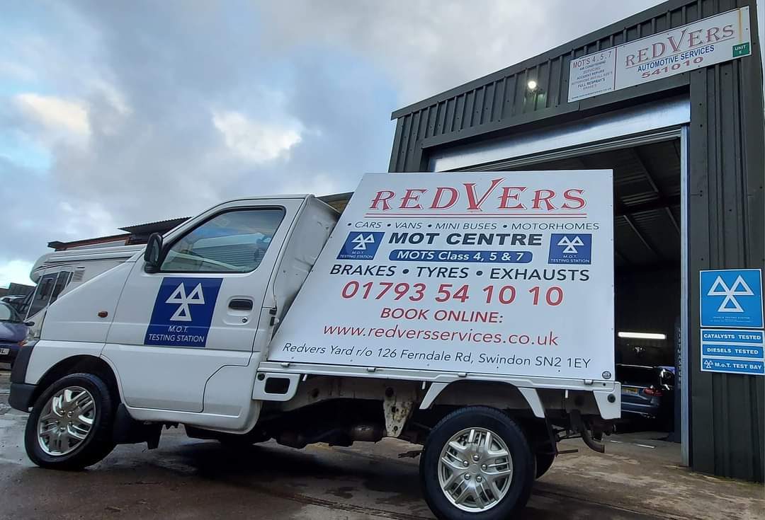 A small white pickup truck with Redvers MOT Centre signage parked outside the garage workshop.