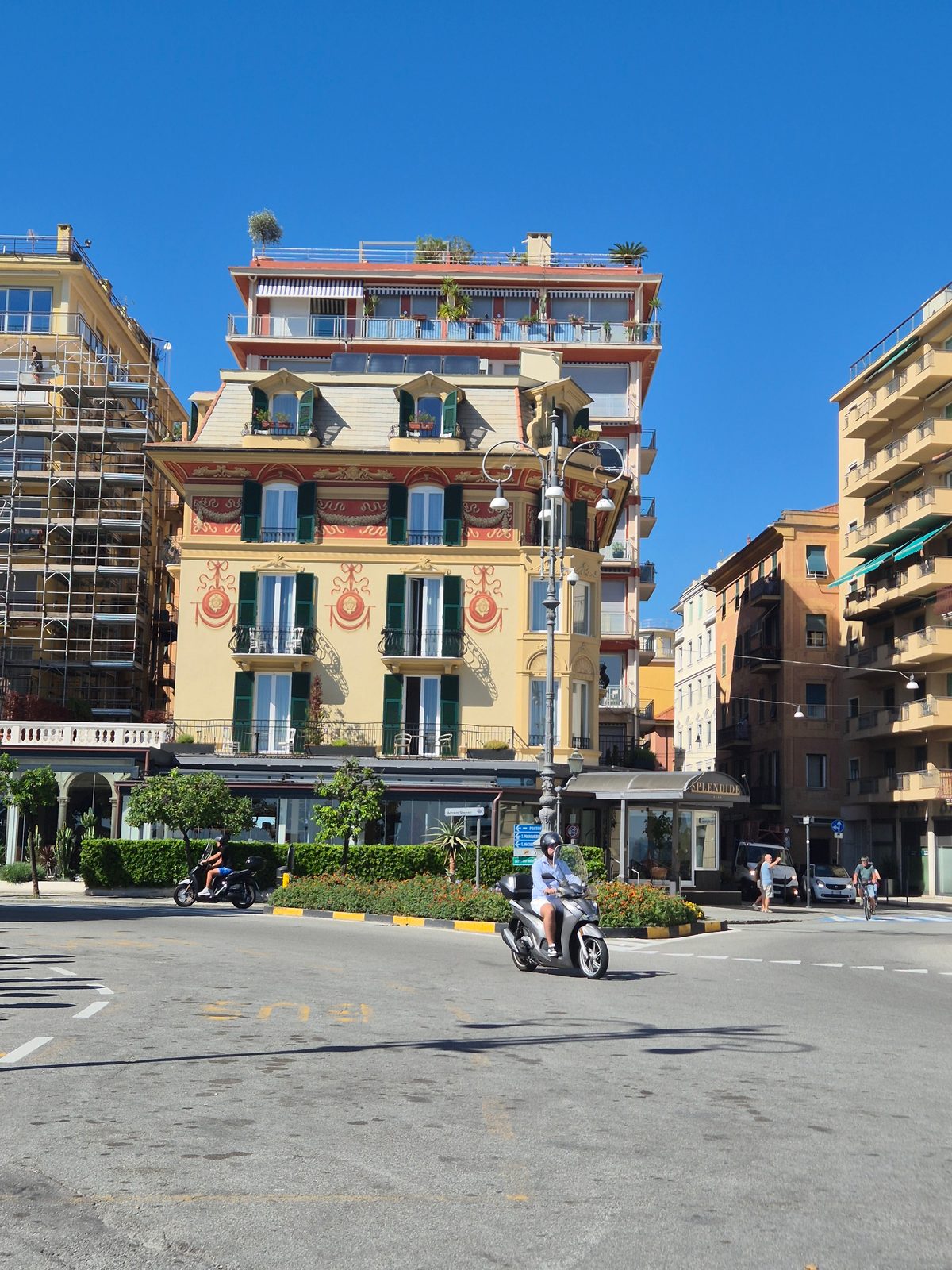 A grand painted Italian building on a street corner under a clear blue sky, with scooters passing through the junction.