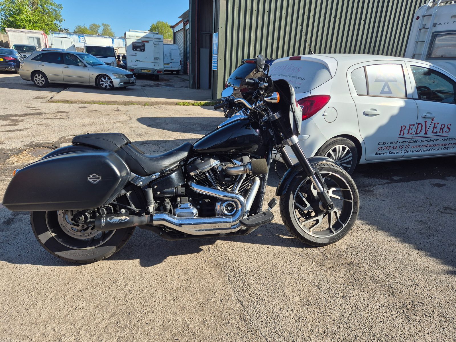 A black Harley-Davidson motorcycle with hard panniers and a windscreen, parked beside a white Redvers branded Vauxhall Corsa, with vans and caravans behind.