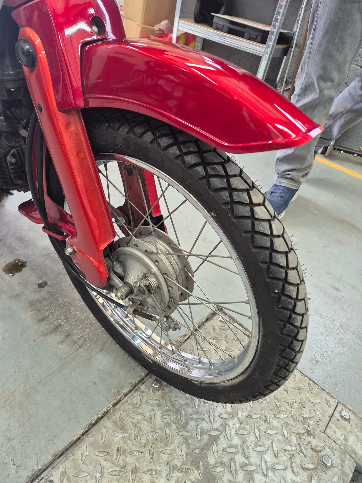 Close-up of a red motorcycle's front wheel with spoked rim, Michelin tyre, drum brake hub and red mudguard, standing on metal chequer plate flooring in a garage.