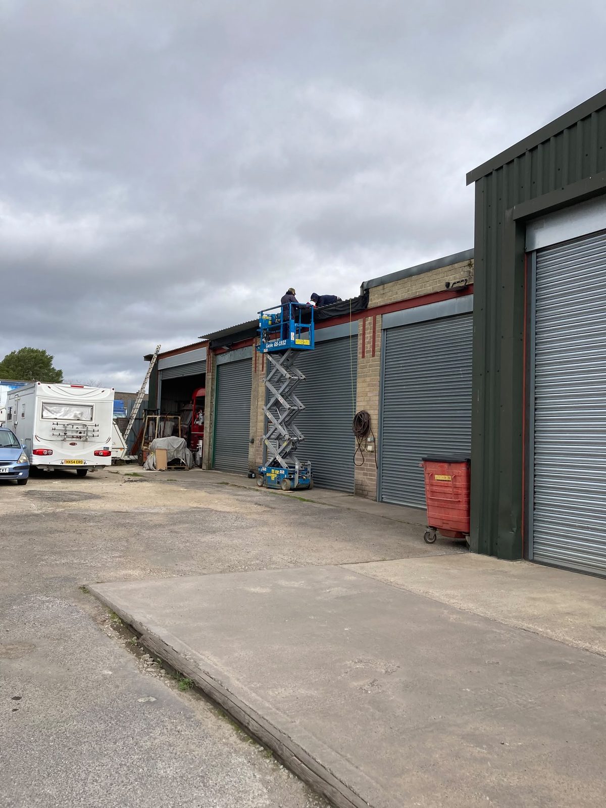 Two workers on a blue scissor lift working on the roof edge of an industrial garage unit with grey roller doors.