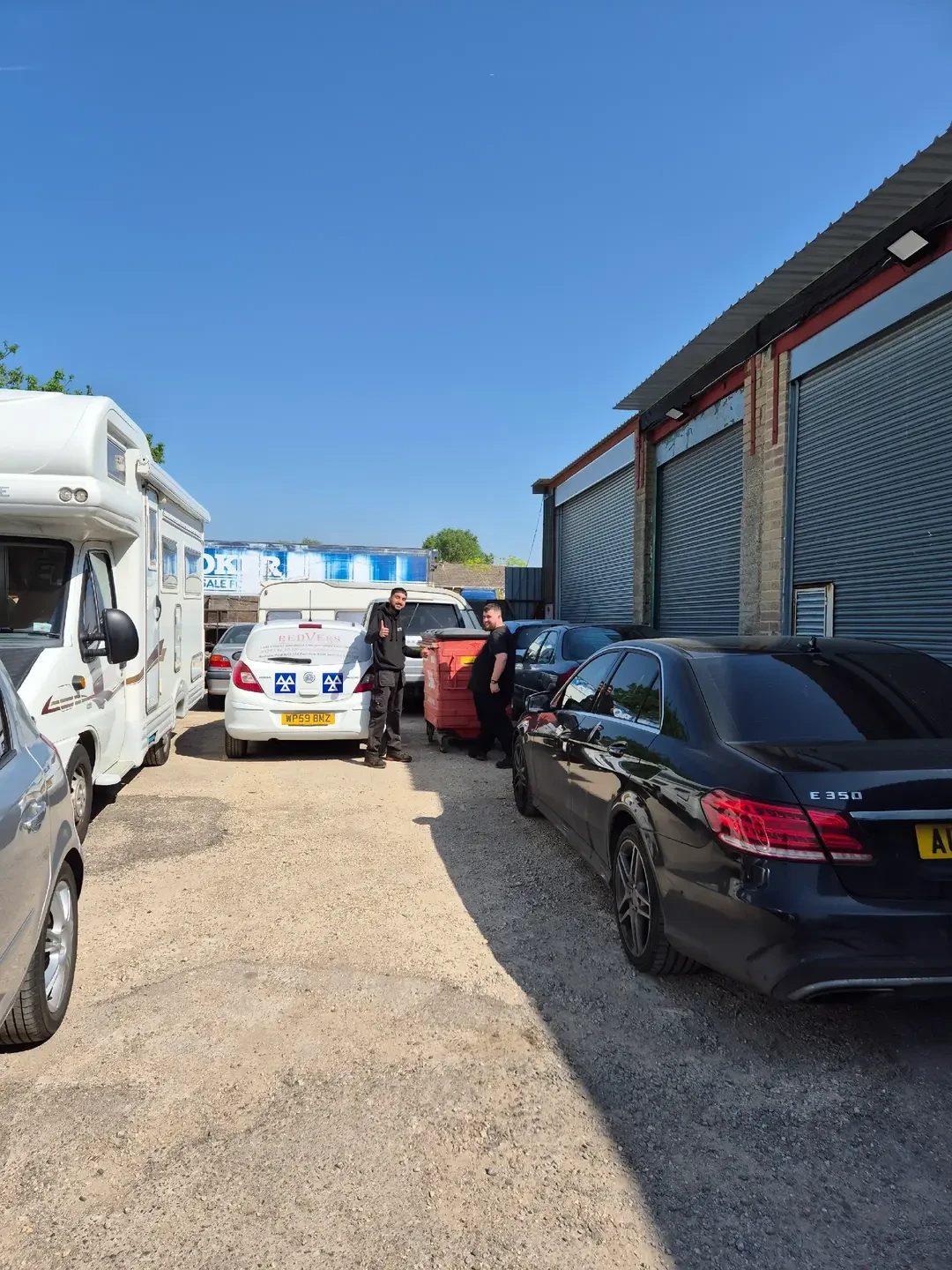 Two men standing on a gravel forecourt beside a white motorhome, a white Redvers van with MOT logos, and a black Mercedes E 350 saloon, next to a unit with grey roller shutters.